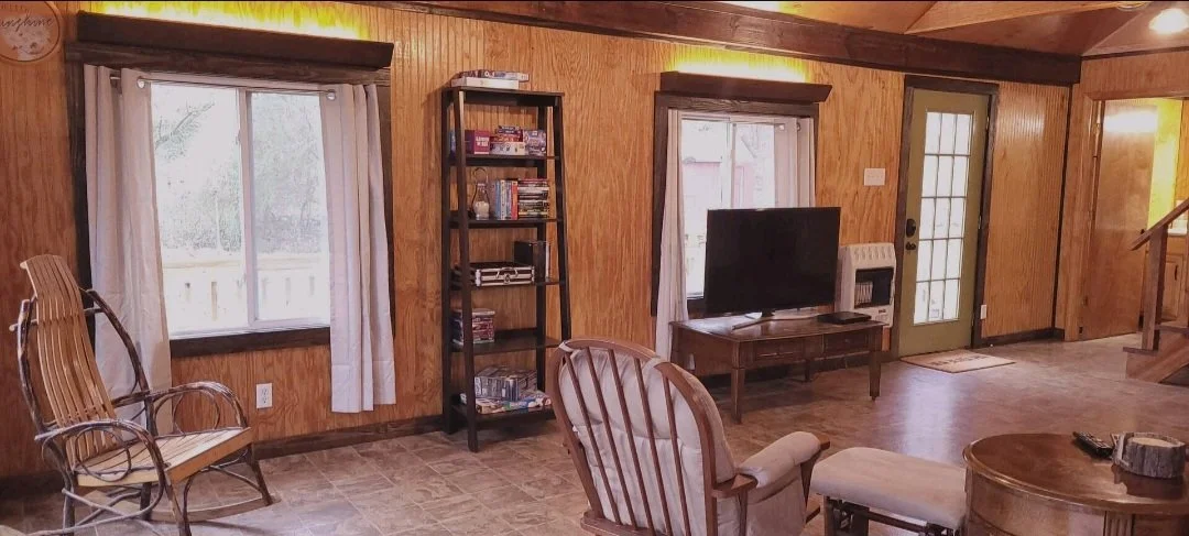 Living room with wooden walls, two windows with white curtains, rocking chairs, a wooden bookshelf with books and electronics, a flat-screen TV on a wooden stand, a door with glass panels, and stairs leading to an upper level.