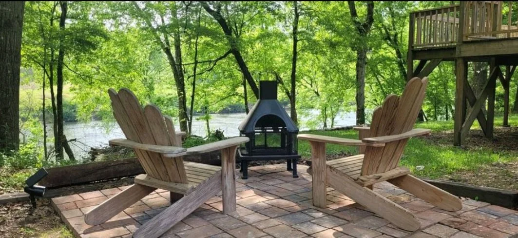 Two wooden Adirondack chairs facing a small outdoor fireplace in a lush green forest near a body of water.