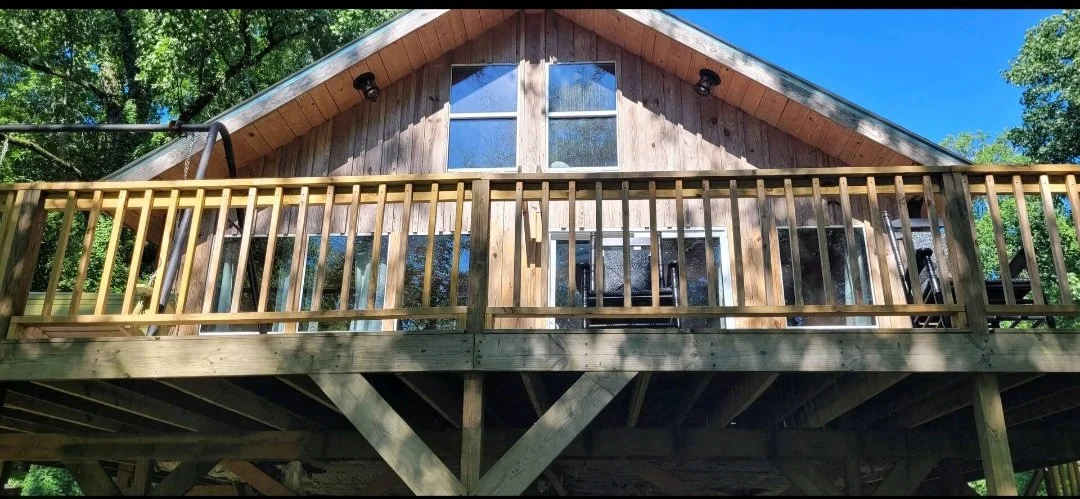 A wooden cabin with a large front window and a balcony, surrounded by green trees under a blue sky.