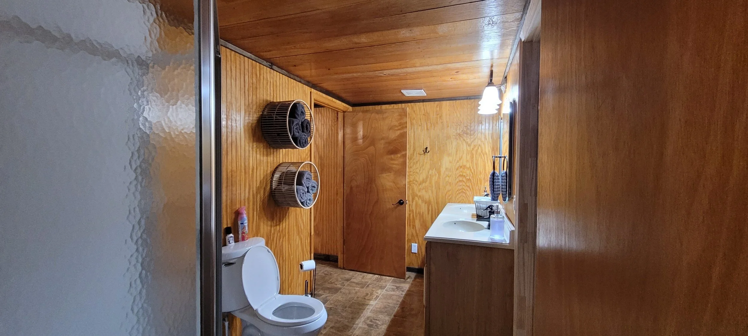 Wood-paneled bathroom with a dual sink vanity, a toilet, and two wicker baskets containing rolled gray towels. The door is closed, and there are hanging towel rings and a light fixture above the vanity.