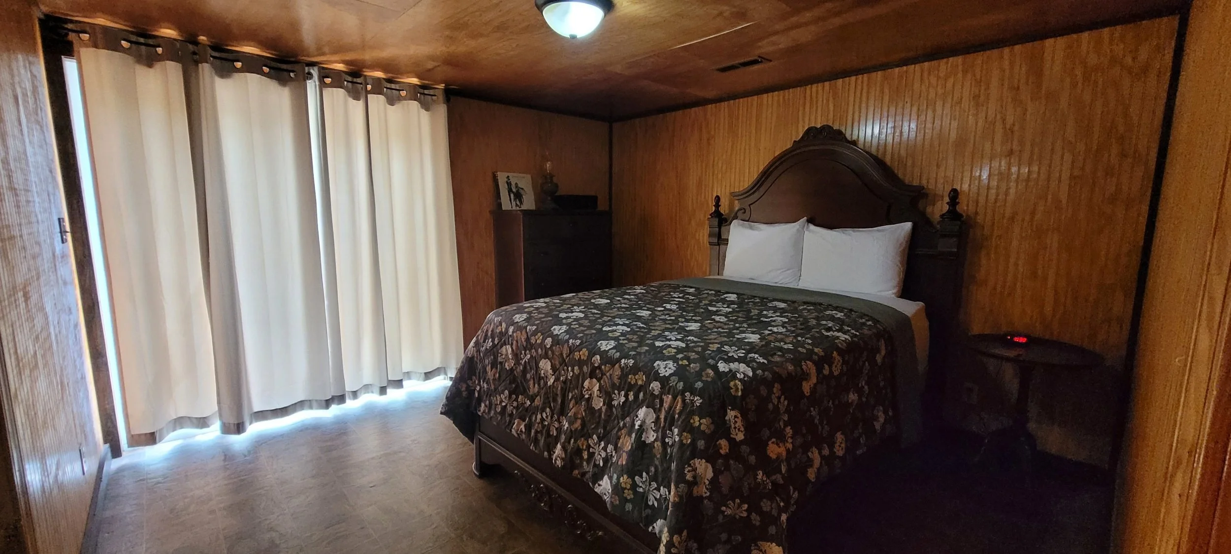 A bedroom with wood-paneled walls, a bed with white pillows and a floral bedspread, a window with light-colored curtains, a nightstand with a clock, and a dresser with framed photo and lamp.
