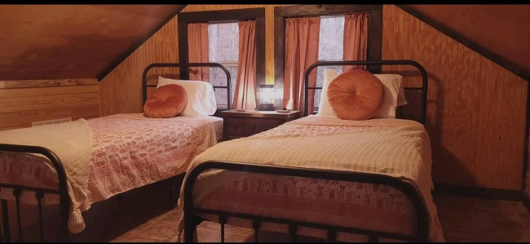 Attic bedroom with two twin beds, pink bedding, round orange pillows, wooden walls, window with pink curtains, small nightstand with lamp in between beds.