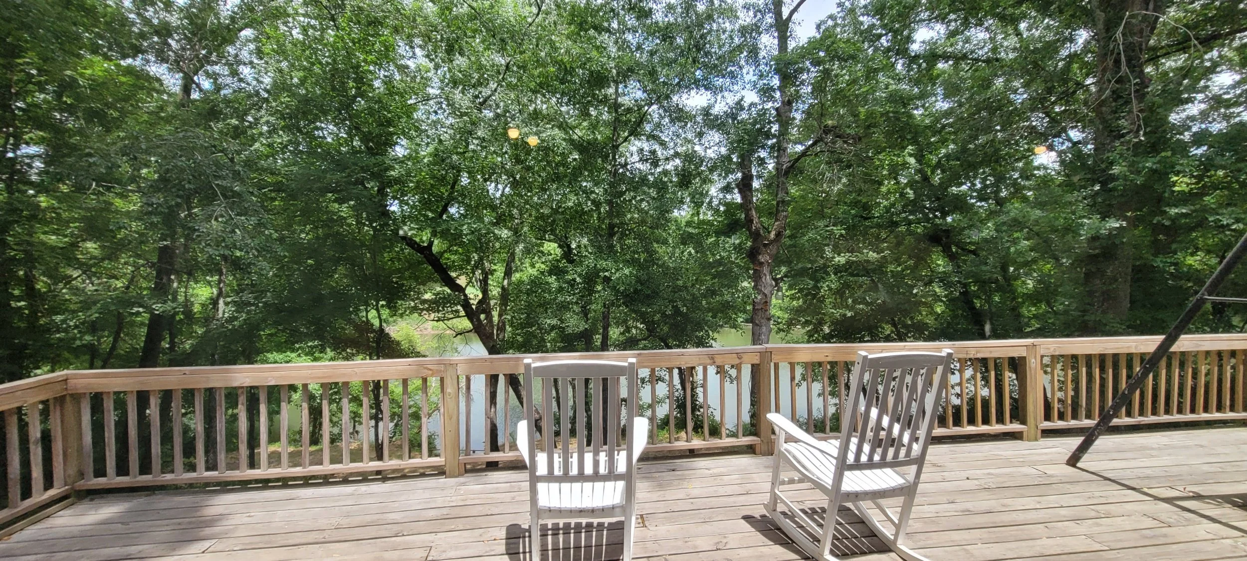 Wooden deck overlooking a lush, green backyard with trees and a pond, featuring a white chair and a white rocking chair.