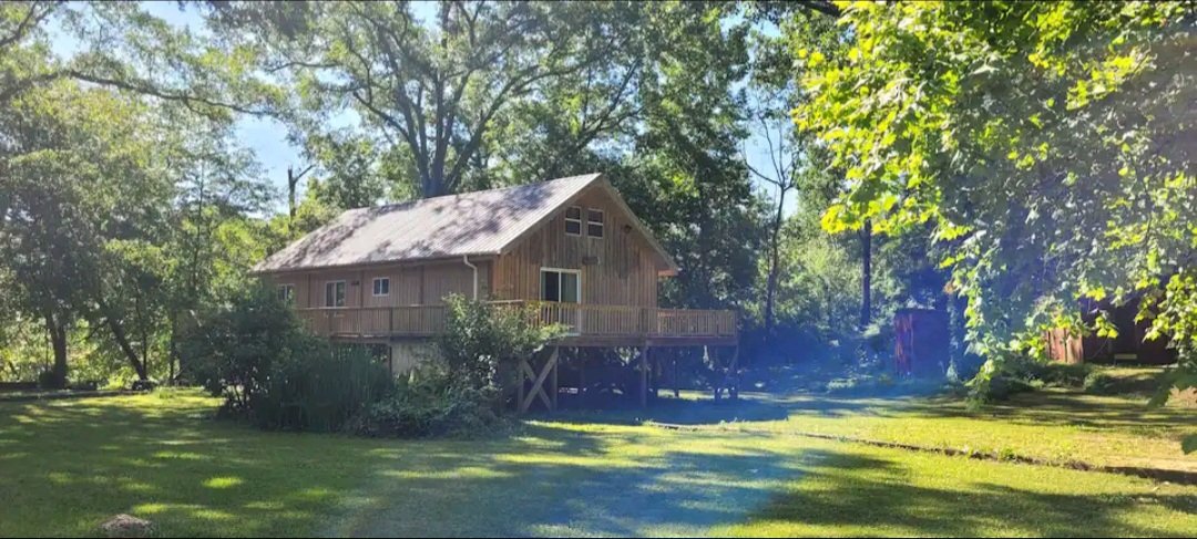A wooden house with a deck, surrounded by green trees and a grassy yard, under a clear blue sky.