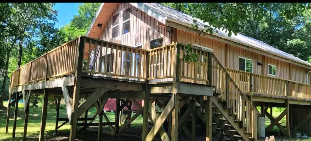 Wooden house with a large elevated deck surrounded by trees in a lush green yard.