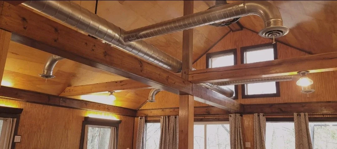 Wood-paneled ceiling with exposed wooden beams, metal HVAC ducts, and a large window with curtains in a cozy room.