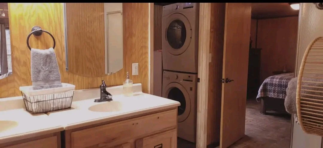 Bathroom with wooden walls, a double sink vanity with a black faucet, a hand soap dispenser, a towel hanging on a ring, and a basket with a towel. In the background, a stacked washer and dryer, and a door leading to a bedroom with a bed.