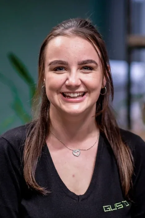 A young women smiling widely at the camera with straight hair. She is in front of a green wall wearing a black jumper that says 'GUSTO'