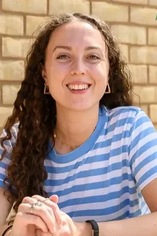 A young women with dark and curly hair sat with her hands together at the table, smiling at the camera. The background is a brick wall with green plants hanging down