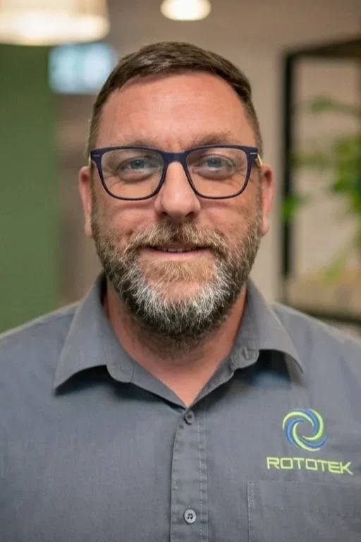A man with black glasses and a stubbled beared smiling in front of a green background with shelving and plants