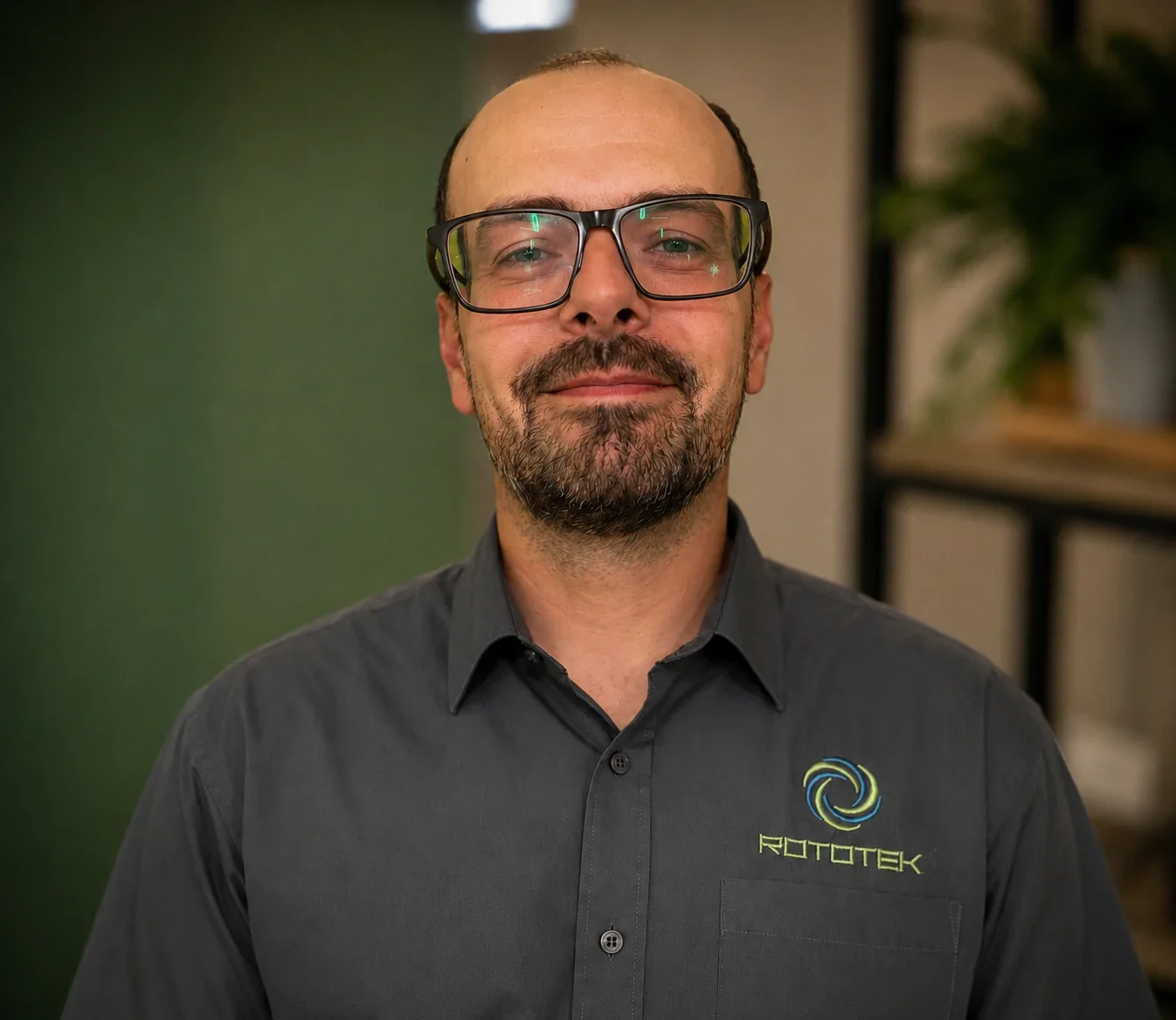 A man with black glasses and a black beard smiling in front of a green background wearing a grey shirt with the words 'Rototek' written on them.