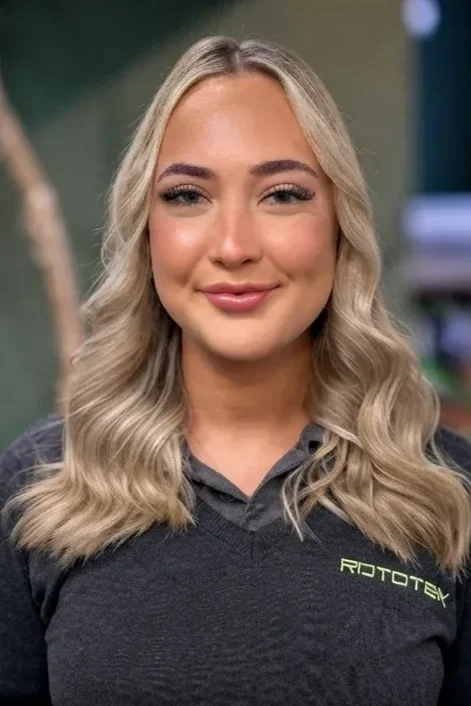 A young lady with blonde curly hair and a Rototek jumper smiling in front of a green wall