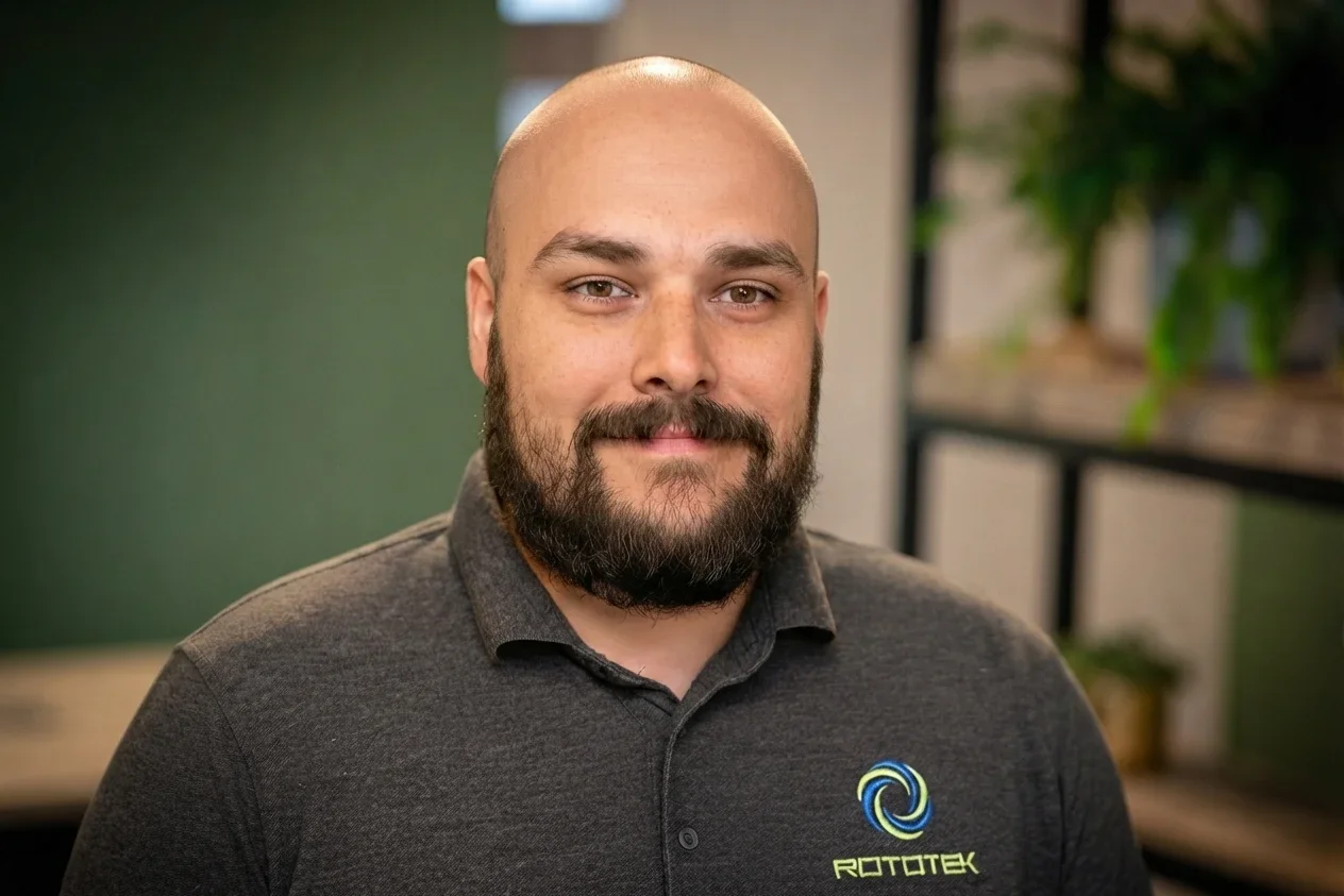 A bald man with a beard and moustache smiling in front of a green background wearing a grey polo shirt that says 'Rototek' on it.