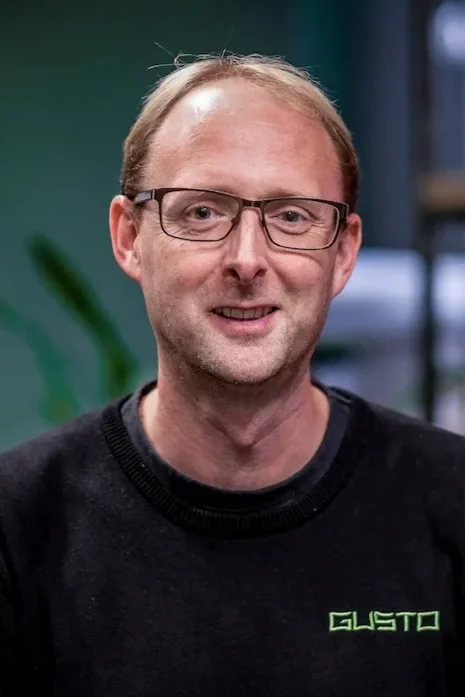A man wearing black glasses and a black jumper that says 'GUSTO'