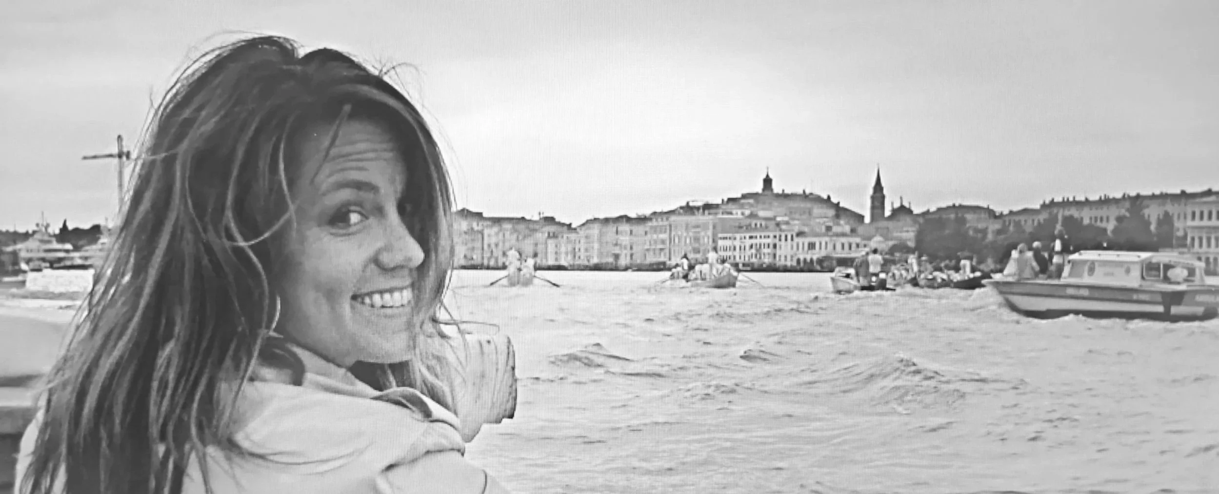 A woman with long hair smiling at the camera on a beach with boats and Venice, Italy in the background, in black and white.