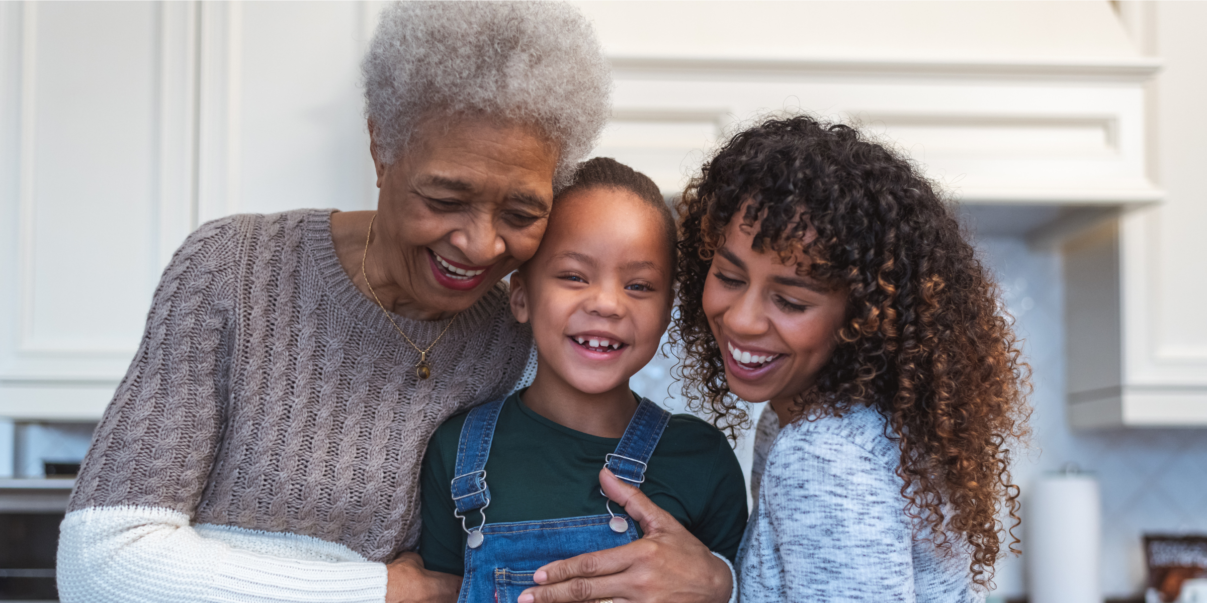 An older woman with short gray hair, a young child in denim overalls, and a younger woman with curly hair all smiling together warmly in a kitchen.
