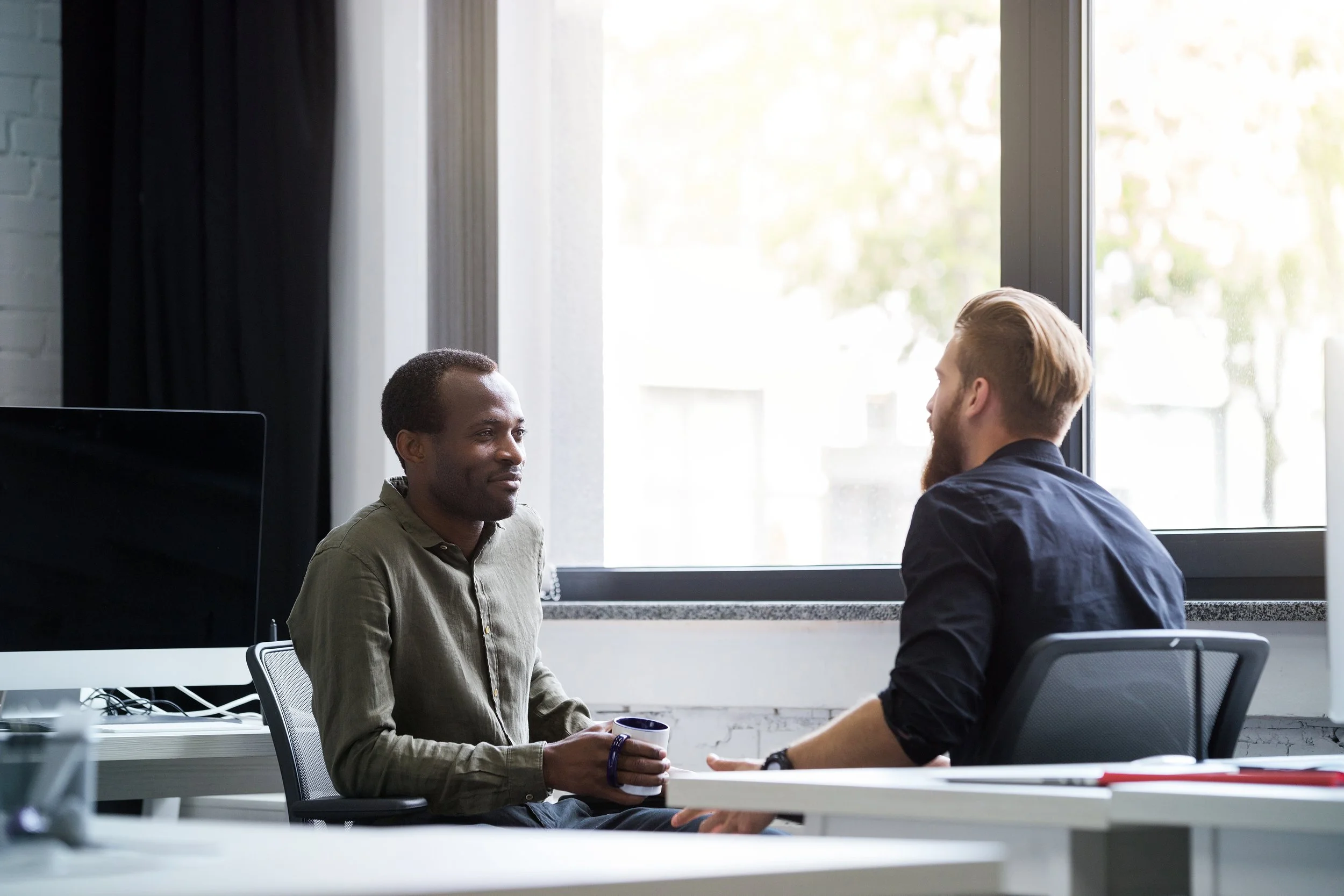 Two persons sitting in an office near a window, having a professional conversation.
