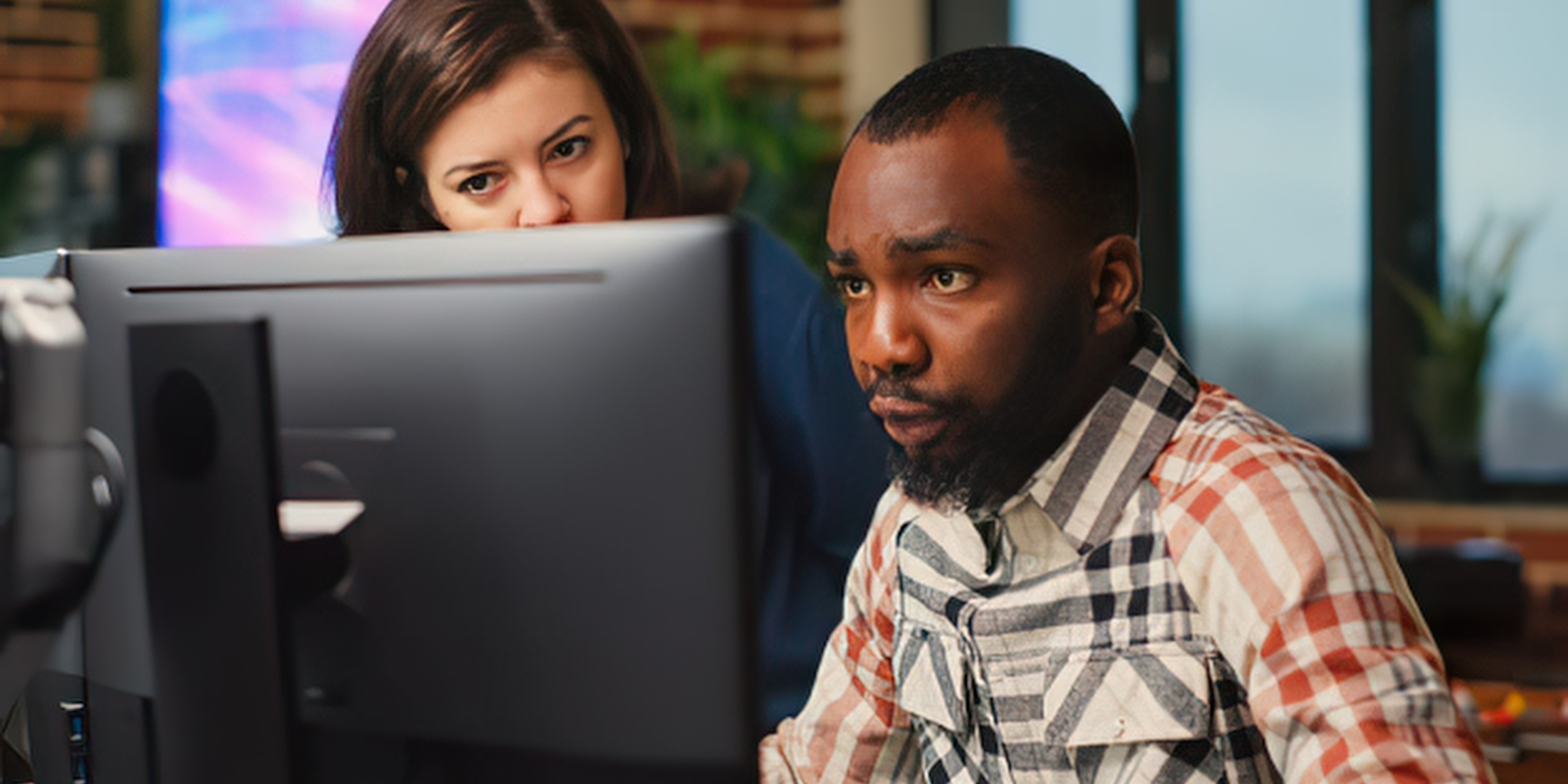 A man wearing a plaid shirt and a woman standing behind him look intently at a computer screen in a modern office setting.