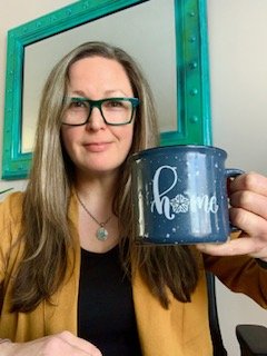 ID: Aimee Whyte, with emerald glasses and light brownish hair, is smiling at the camera. She is holding a "home" mug (with the 'o' in home depicting the flour/flower symbol for Rochester, NY). A bright turquoise rectangular mirror hangs behind her. S