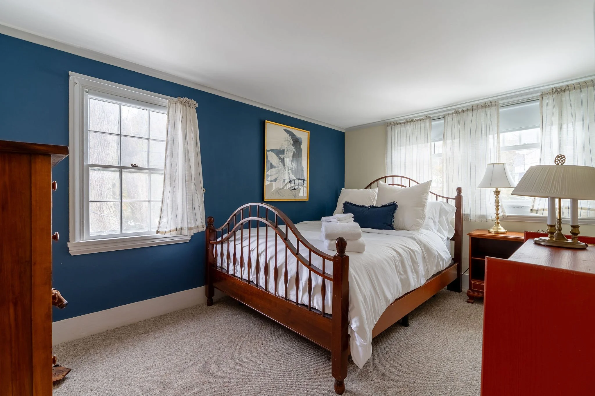 A cozy bedroom with a wooden bed frame, white bedding, and pillows. There are nightstands with lamps on either side and large windows with sheer curtains, allowing natural light in. A framed artwork hangs on the teal accent wall.