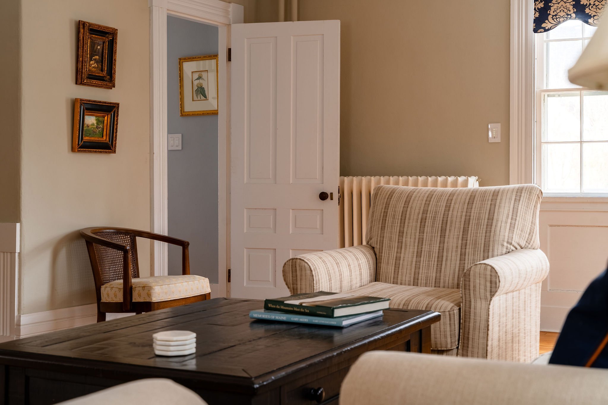 A cozy living room with a plaid armchair, a wooden coffee table with books on top, a side chair with a cane back and cushioned seat, and a window with a patterned valance. There are framed pictures on the wall near the door and a radiator behind the 