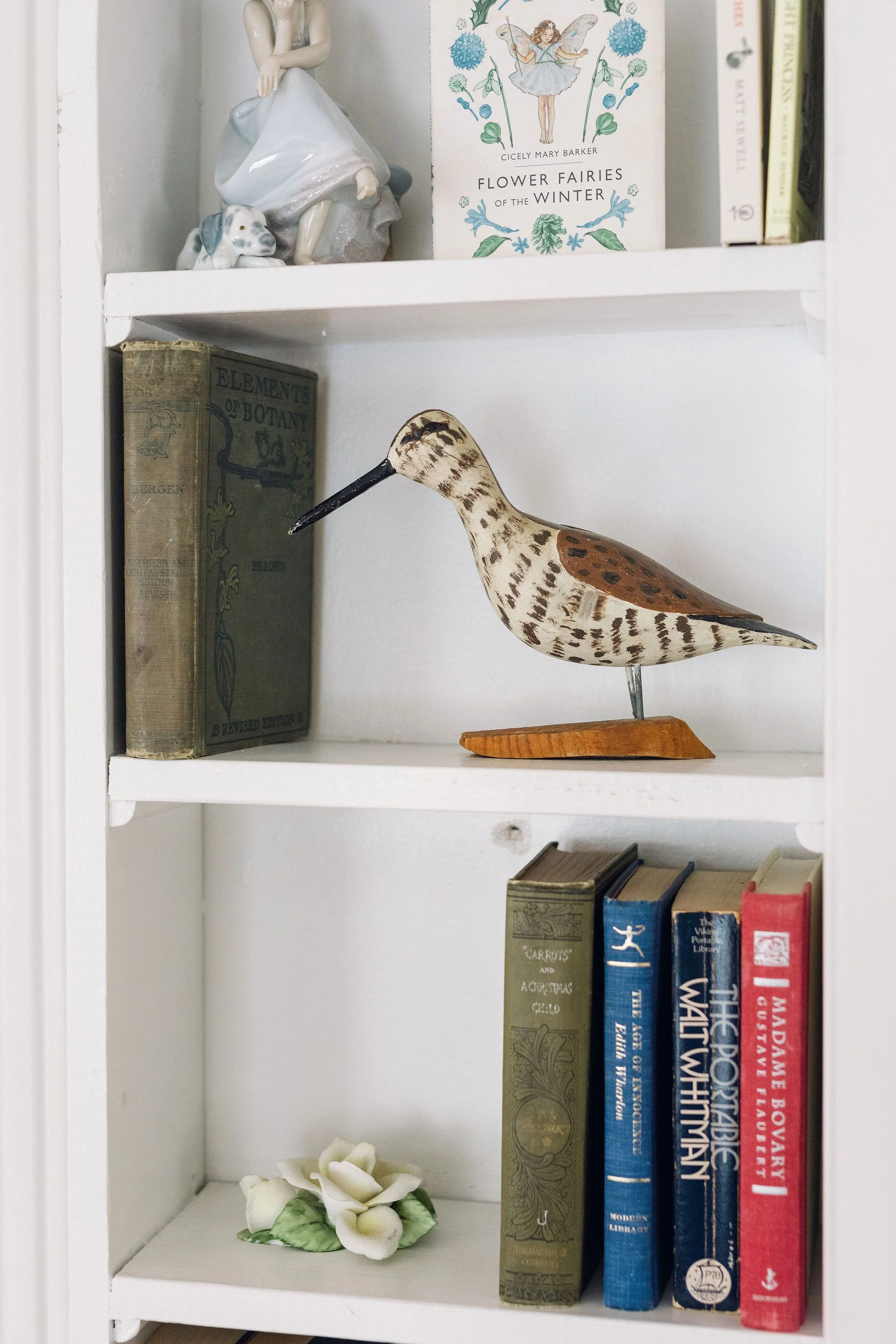 A white bookshelf with decorative items and books, including a bird figurine, a ceramic girl with a dog, and a small porcelain flower, arranged on three shelves.