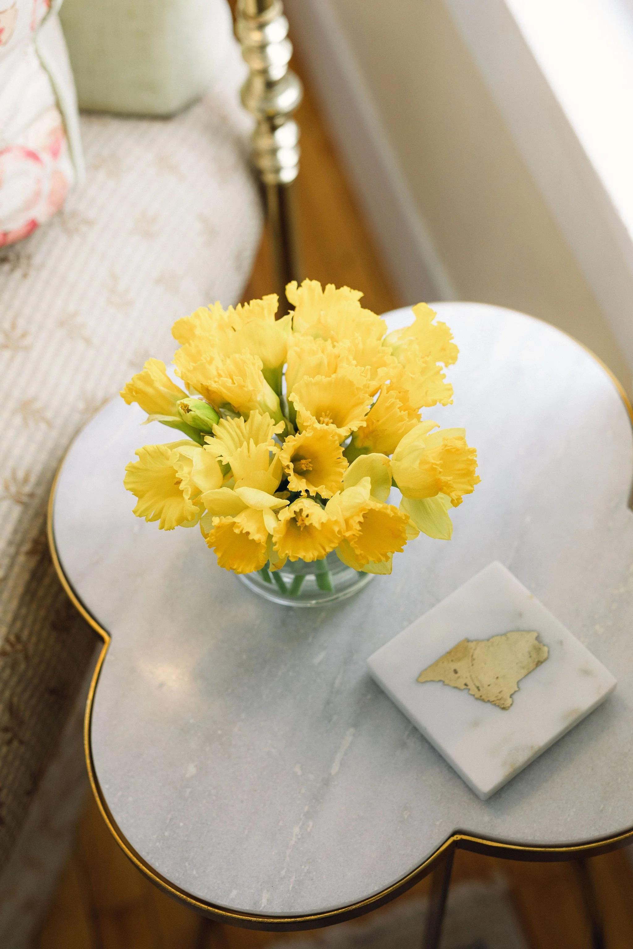 Yellow daffodil flowers in a glass vase on a white marble side table with a piece of gold leaf decor on a white marble coaster.