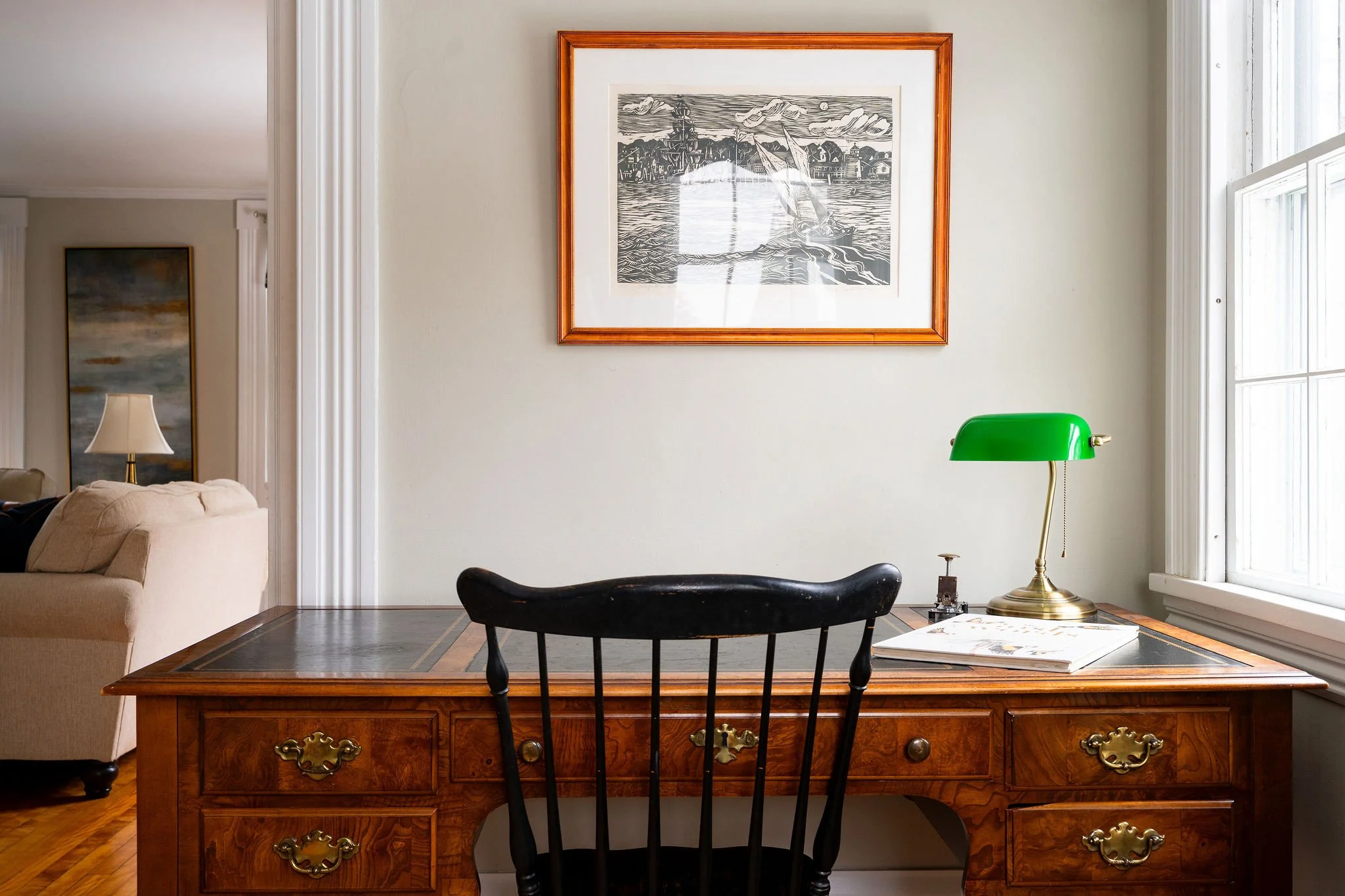 Wooden desk with a green banker’s lamp, a black chair, a framed black-and-white artwork on the wall, and a window to the right letting in natural light.