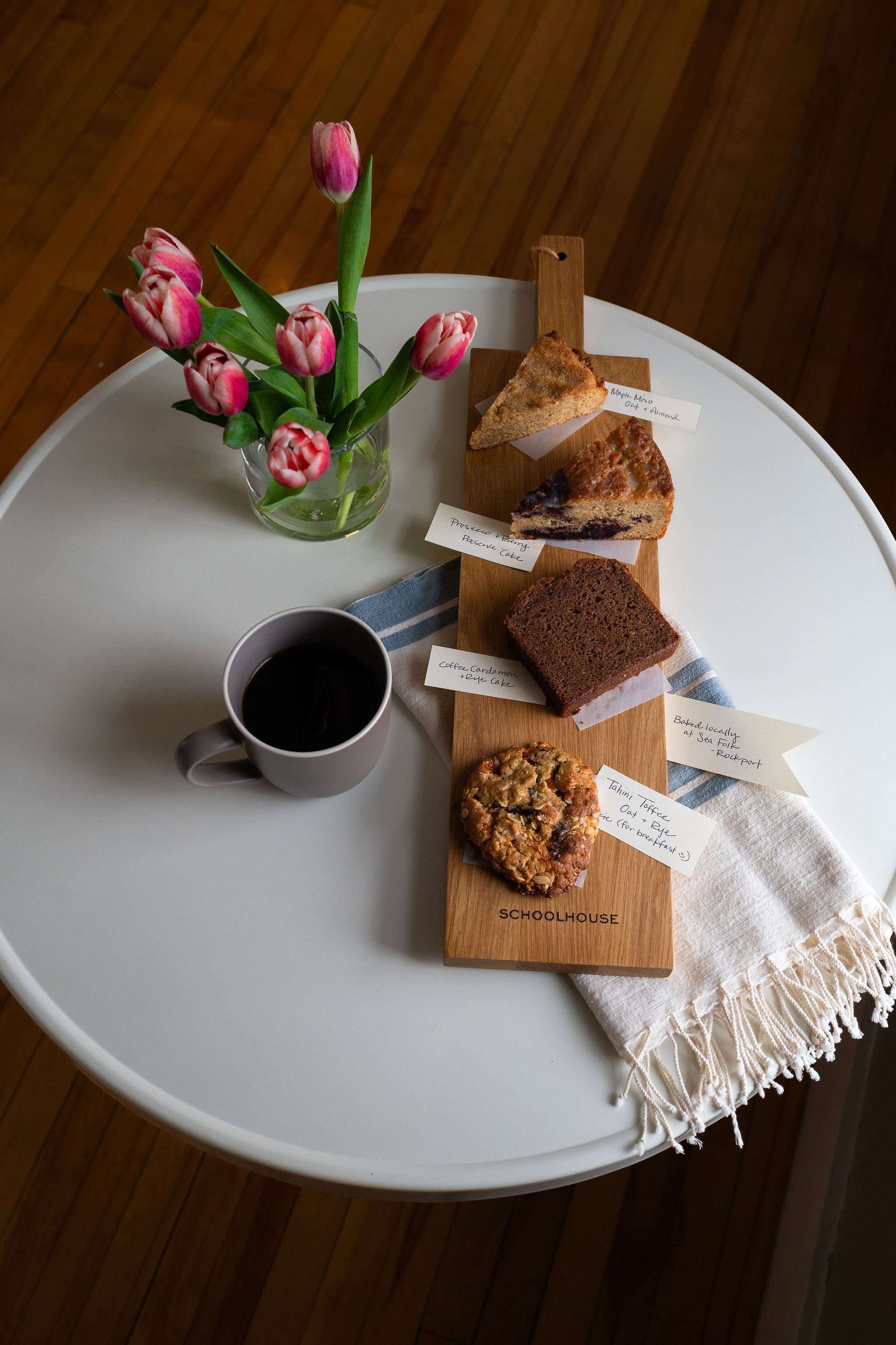 A table with a vase of pink tulips, a cup of black coffee, and a wooden serving board with various slices of cake, each labeled with handwritten notes. The table has a white surface, and the background features wooden flooring.