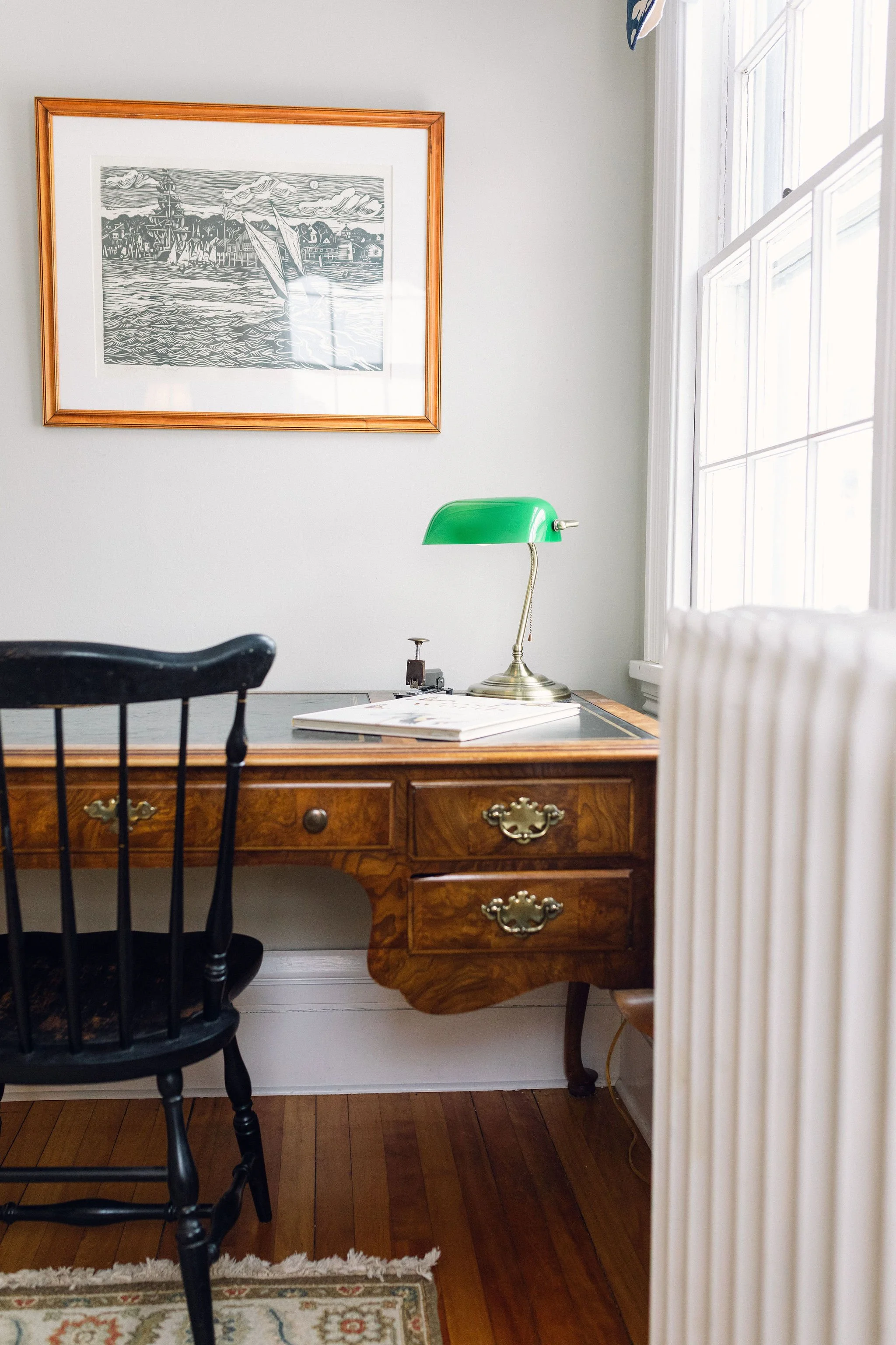 A vintage wooden desk with brass handles, green banker style desk lamp, black wooden chair, framed black and white artwork, window with white framing, hardwood floor, and a small patterned rug.