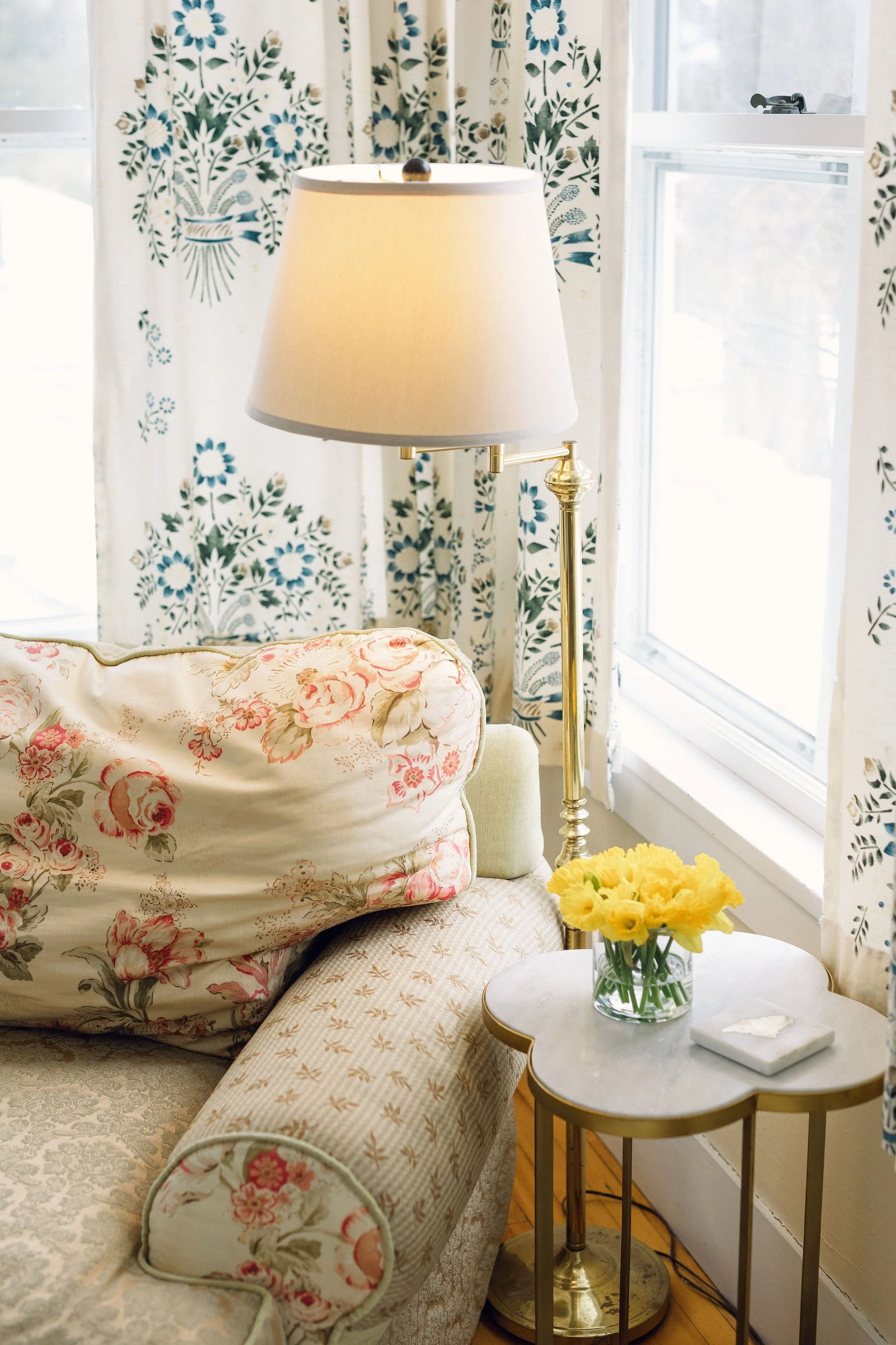 A living room corner with floral-patterned curtains, a table lamp with a beige lampshade, a floral cushion on a vintage sofa, a round side table with a vase of yellow flowers, and a window with white trim.