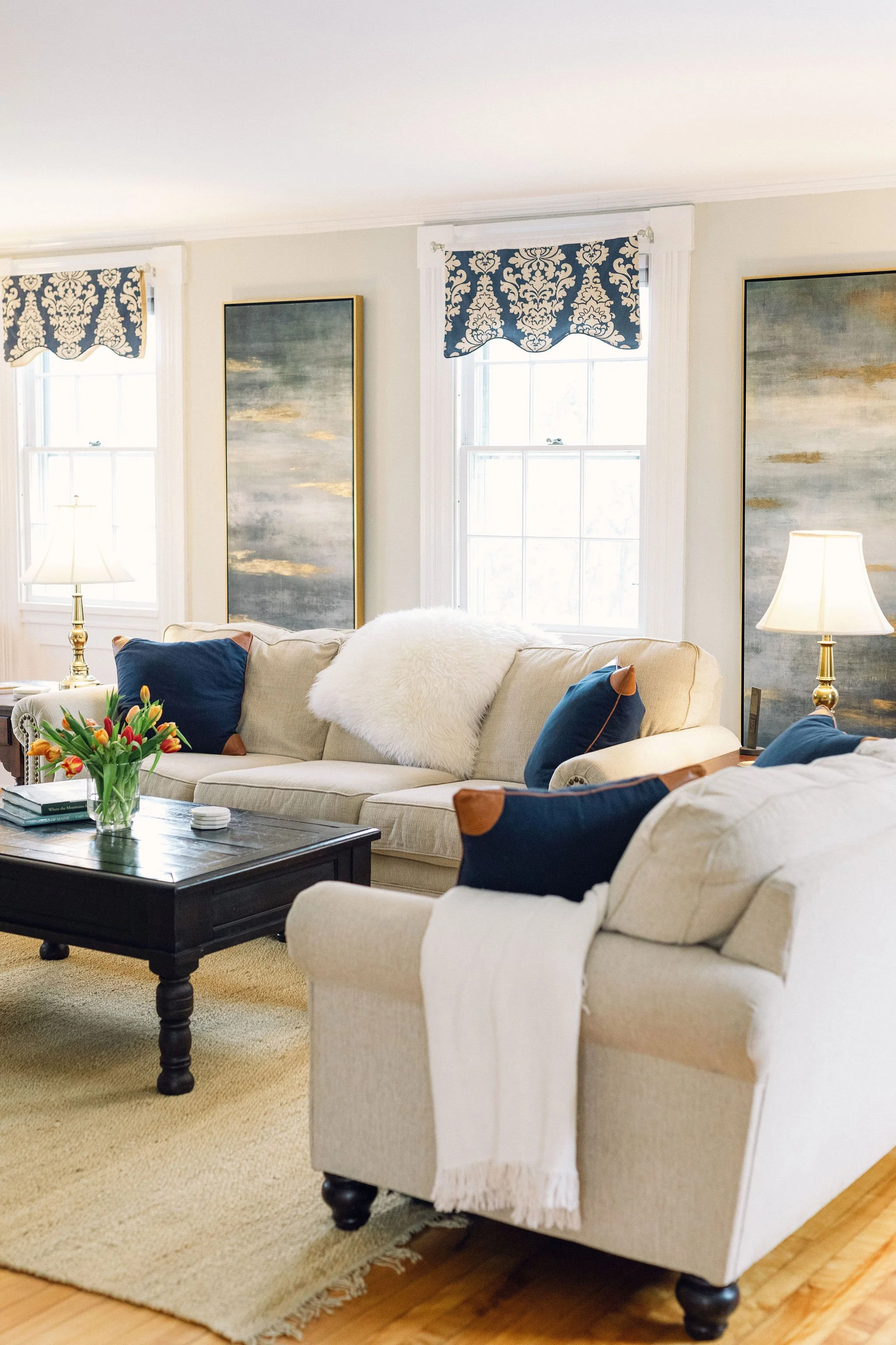 Bright living room with beige sofas, navy pillows, a coffee table with a vase of tulips, side lamps, and wall art with blue and gold hues, featuring two windows with valances.