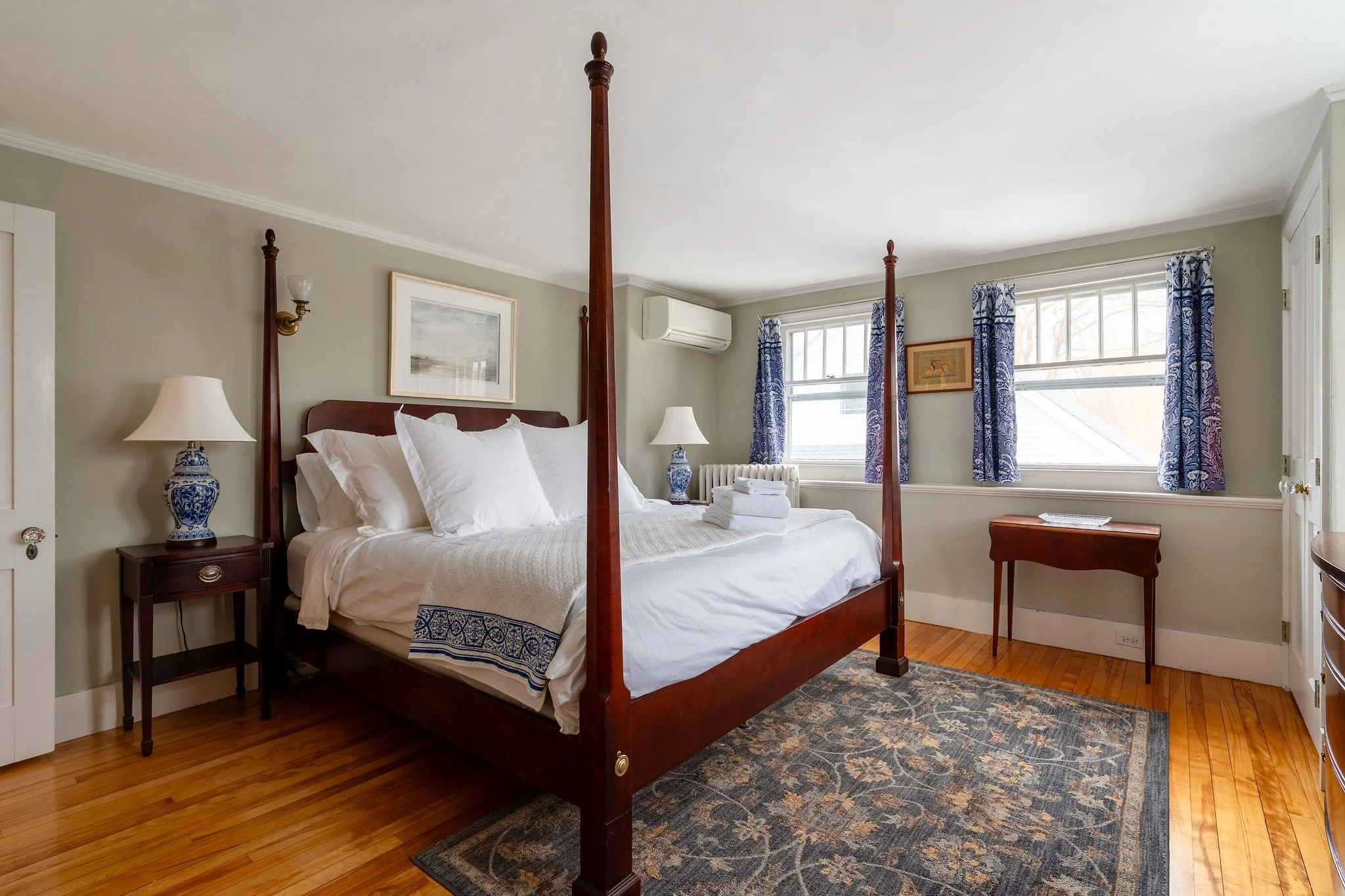 A bedroom with a four-poster wooden bed, white bedding, blue and white lamps, framed artwork, and windows with blue curtains.