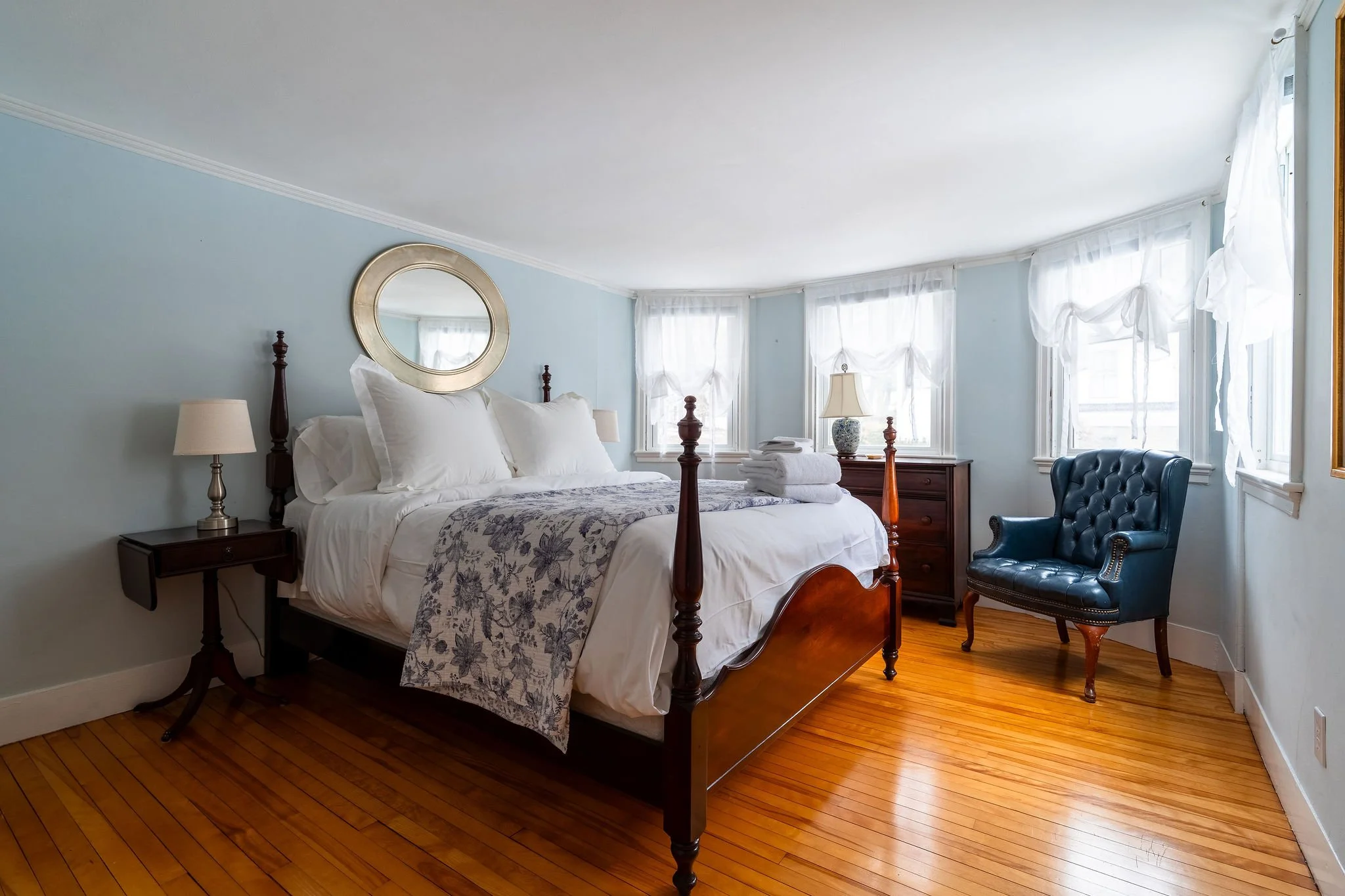 A bedroom with a wooden four-poster bed, white linens and a floral bedspread, two bedside tables with lamps, a dresser with a lamp and towels, a blue tufted chair, and a large mirror with white curtains on multiple windows, hardwood floor, and light blue walls.