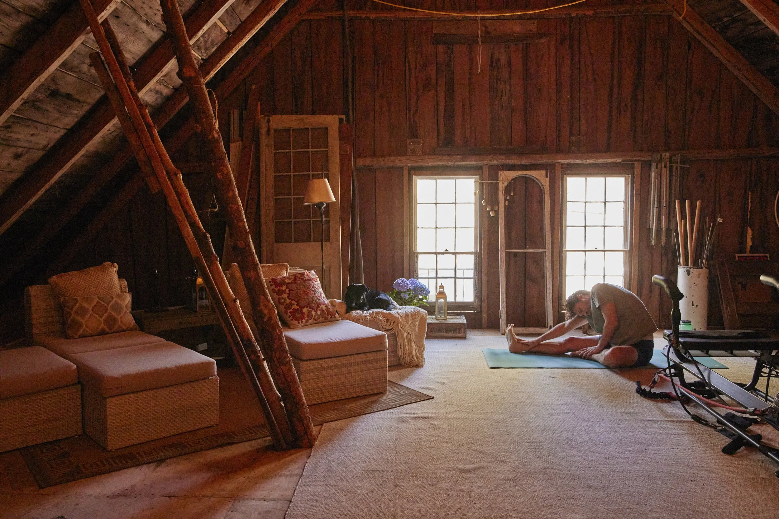 An interior view of a rustic wooden attic or barn workspace with two windows, a treadmill, a sofa with pillows, and various vintage and industrial decor items.