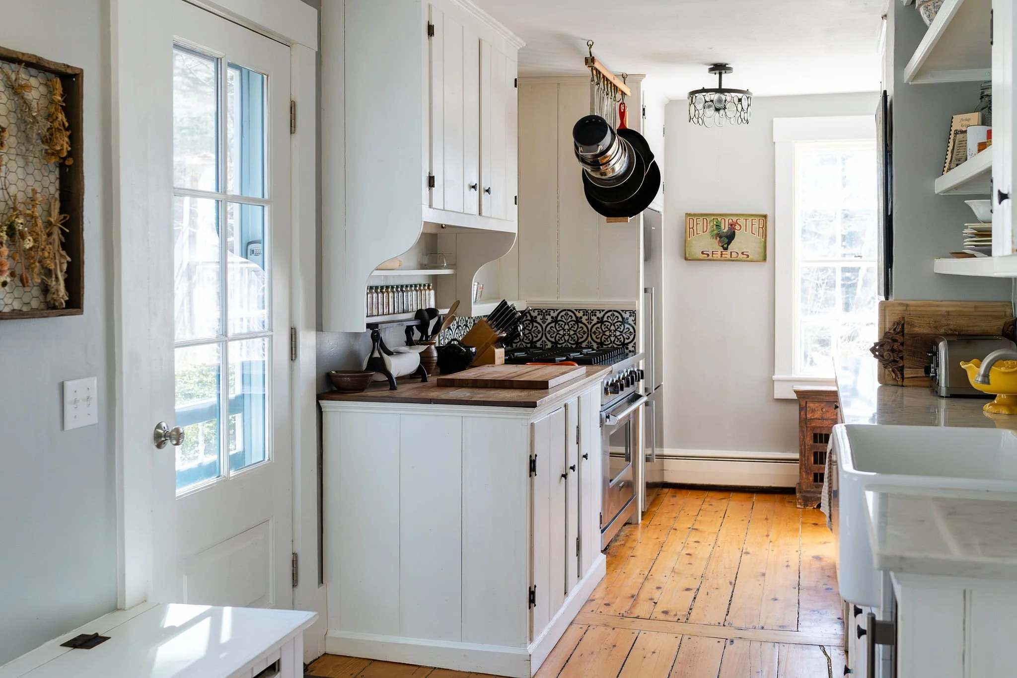A cozy kitchen with wooden open shelving, black cast iron skillets hanging, a red kettle, toaster, microwave, and refrigerator. There is a wooden bowl with a rolling pin on a counter, and a large pendant light hanging from the ceiling.