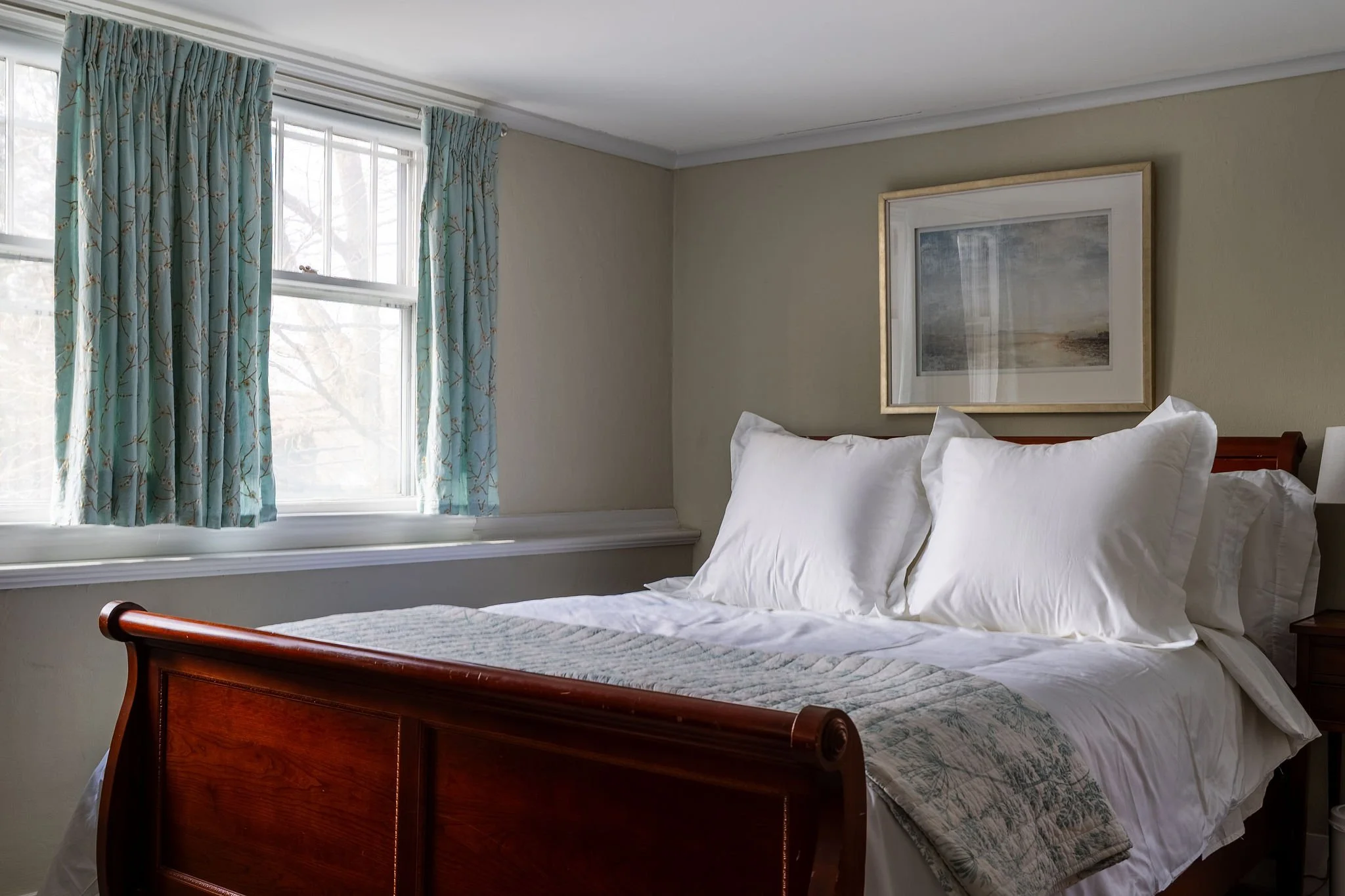 A bedroom with a wooden bed frame, white bedding, and a quilted blanket, next to a window with blue floral curtains and a framed picture on the wall.