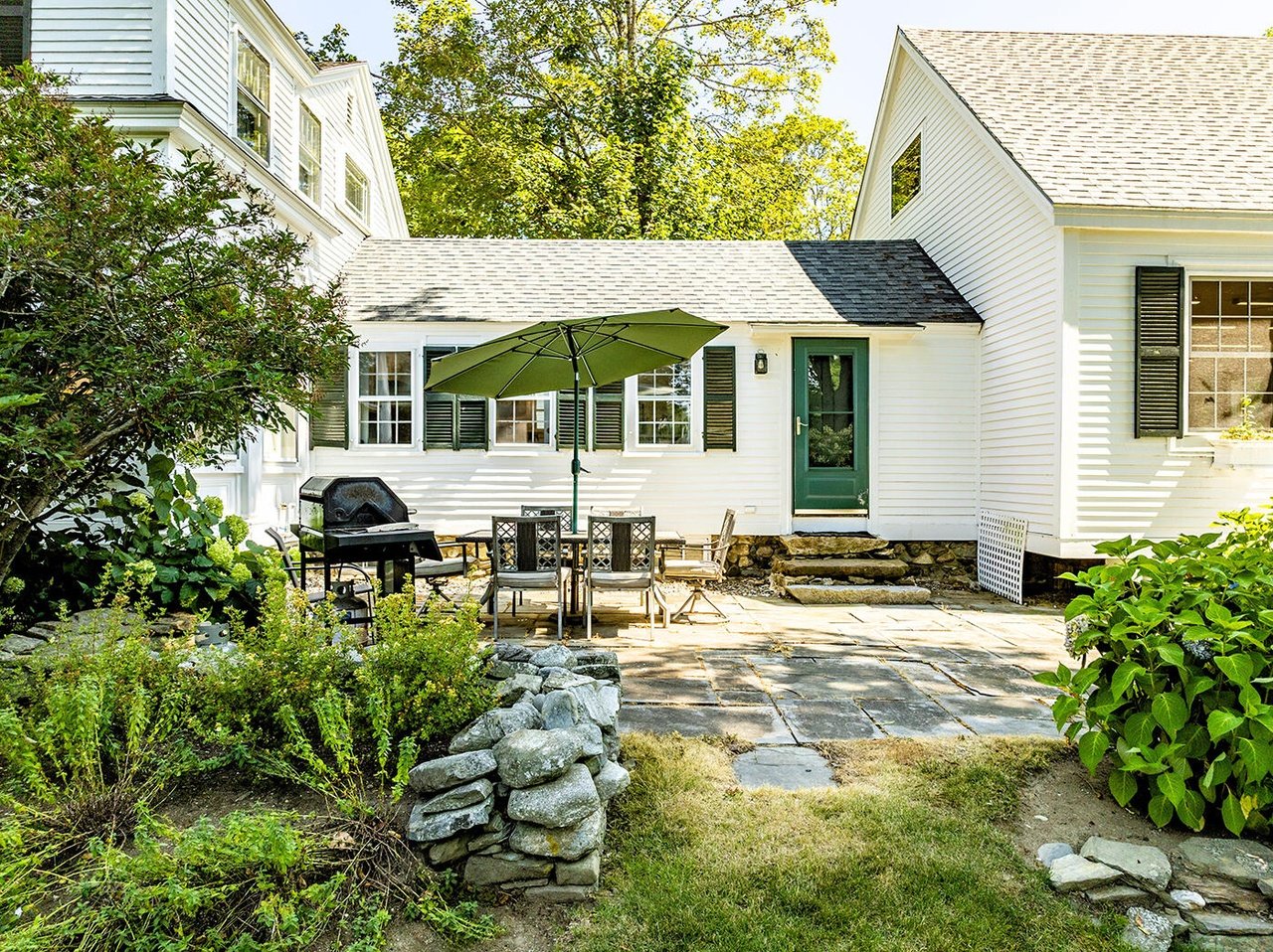 Backyard patio with outdoor dining set, green umbrella, barbecue grill, plants, stone pathway, and a white house with green door and black shutters.