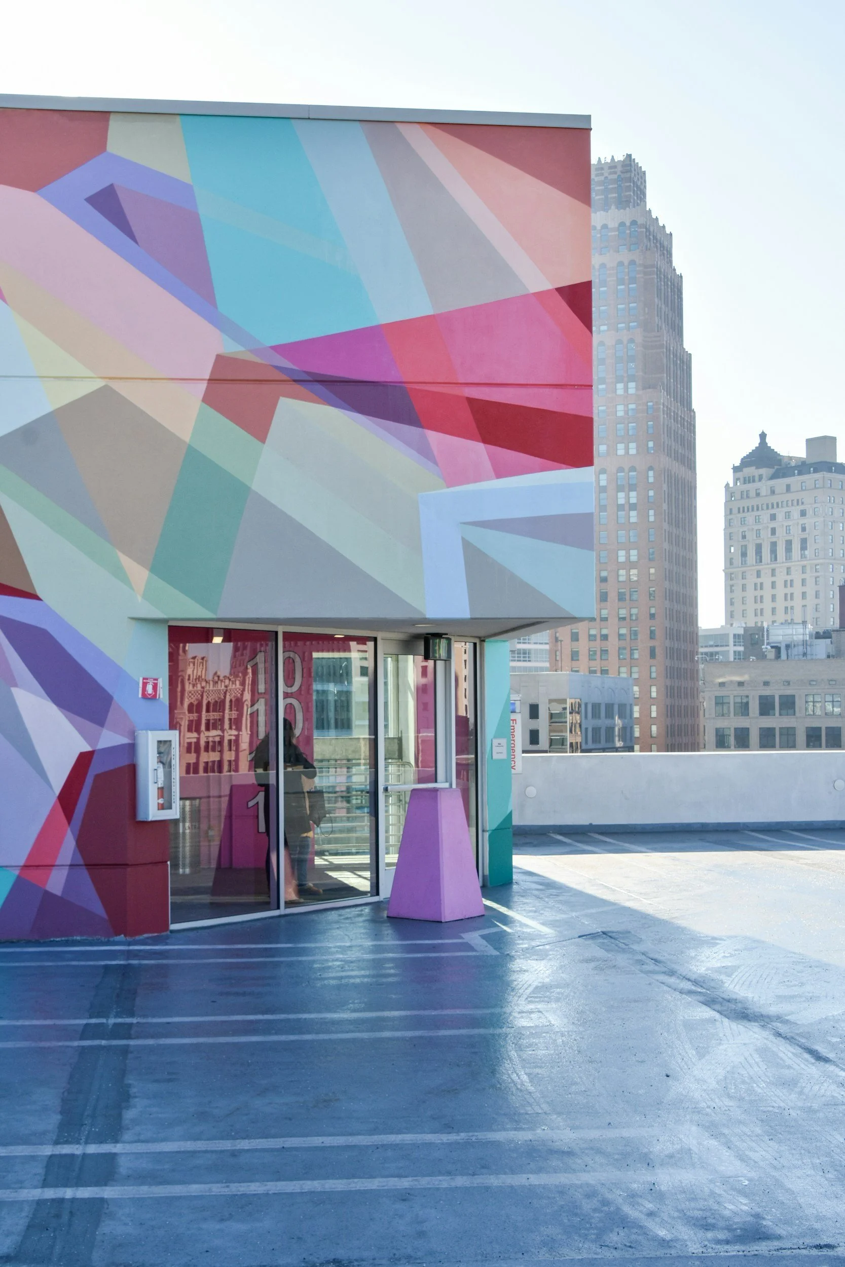 Colorful geometric mural on the exterior wall of a building with a glass door, located in an urban setting with skyscrapers in the background.
