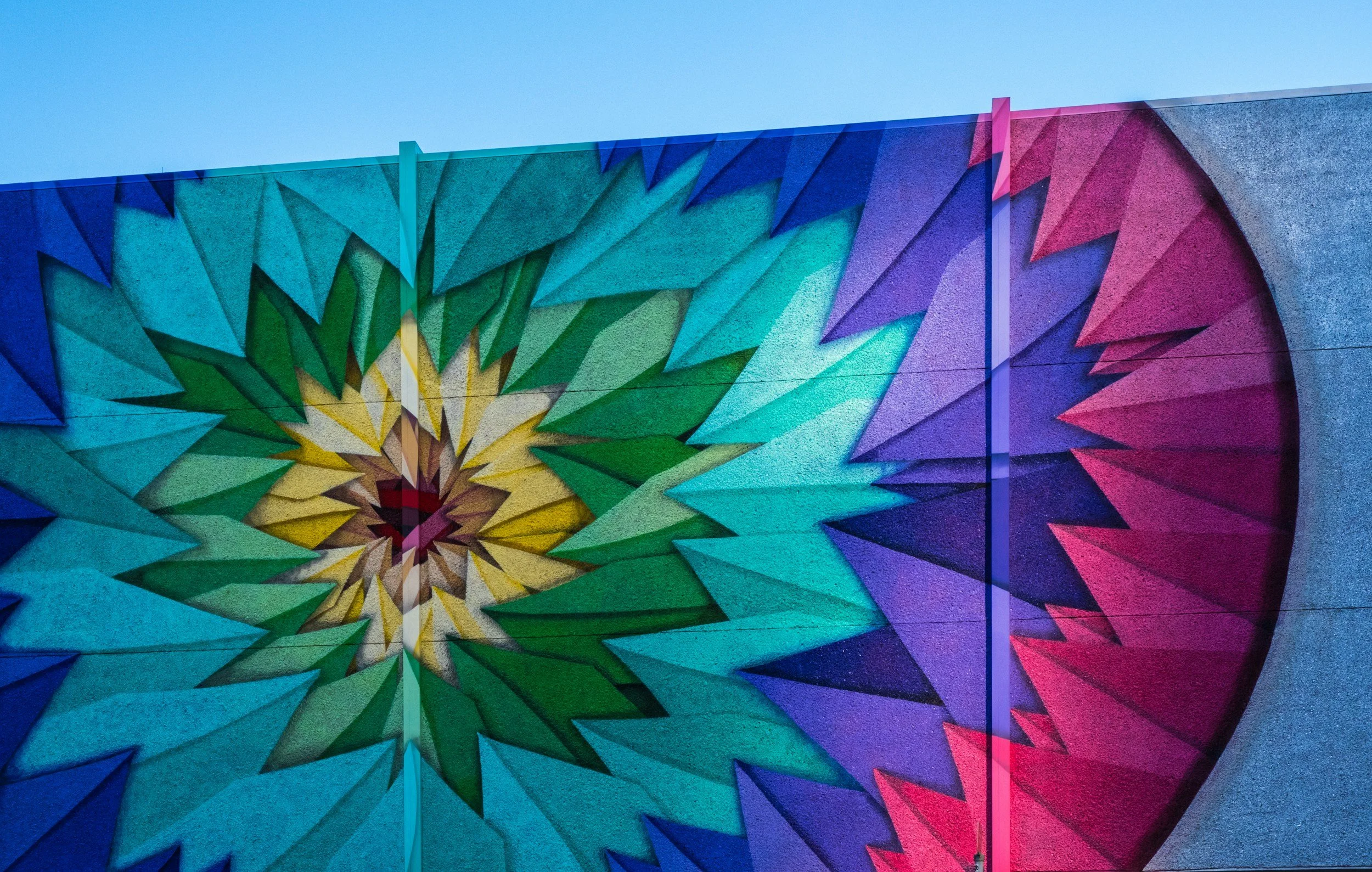 Colorful abstract mural with geometric shapes resembling a starburst pattern in vibrant shades of blue, green, yellow, violet, and pink.