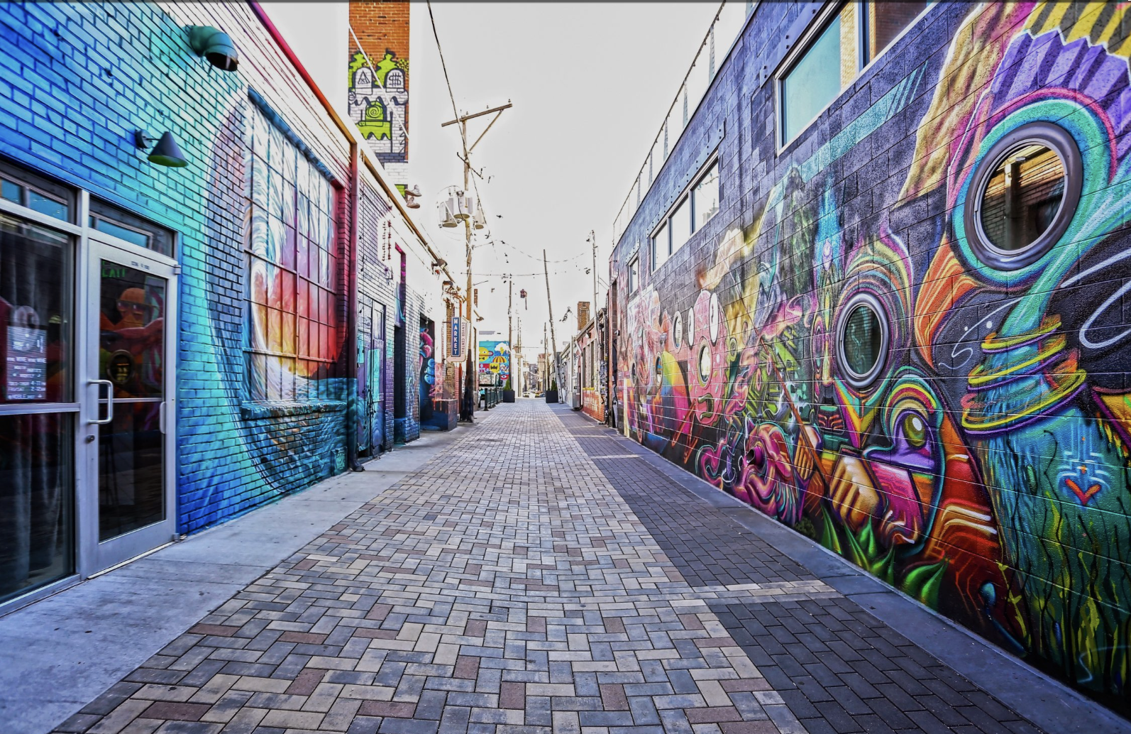 A colorful alleyway with vibrant street art murals on brick walls on both sides, featuring abstract and psychedelic designs, with a paved walkway and power lines overhead.