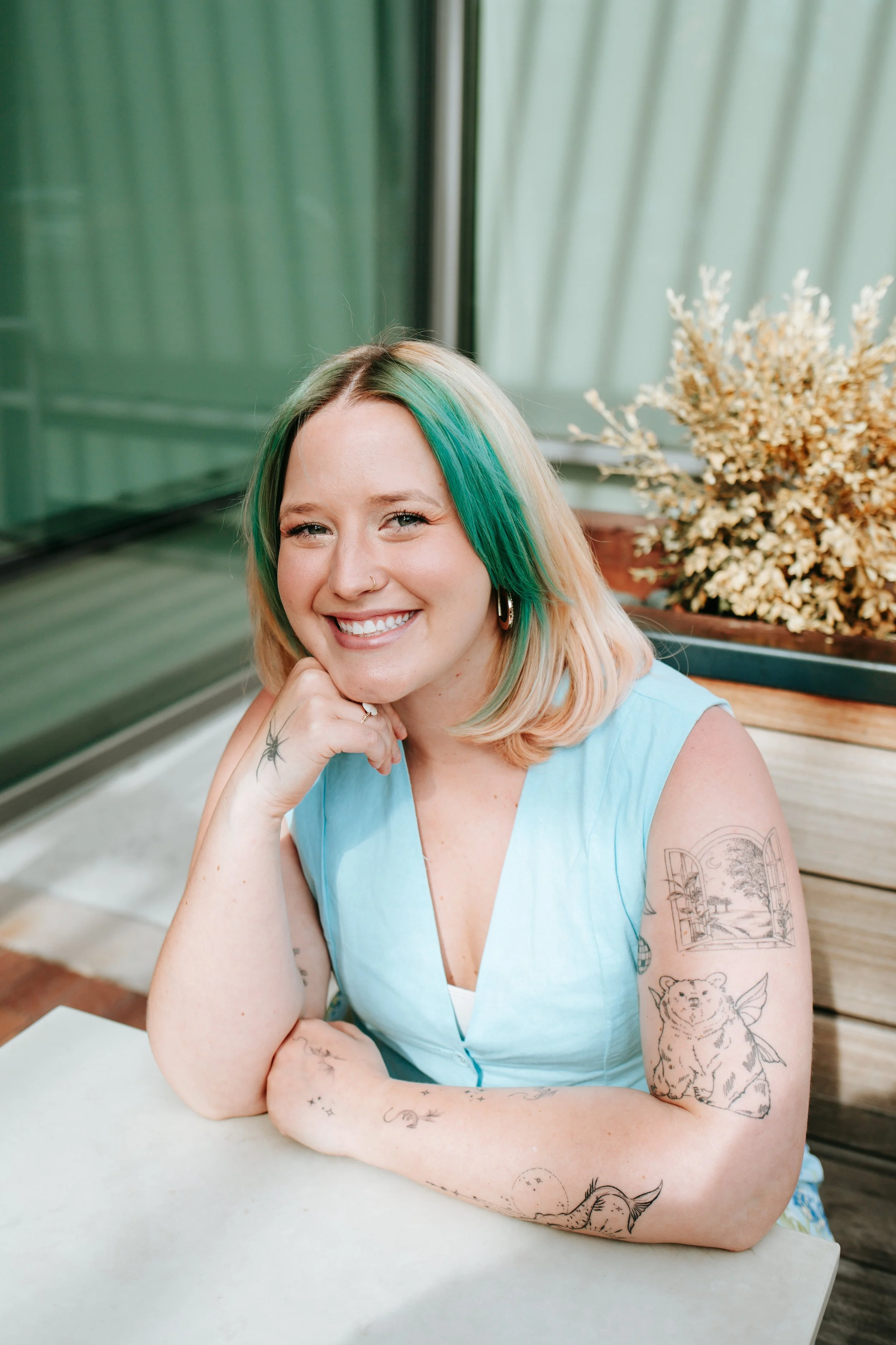 A woman with long, wavy, pinkish-blonde hair, smiling, wearing a black top with sheer patterned sleeves, a silver necklace, and a ring with a green stone. She has a tattoo of a spider on her hand and a nose ring.