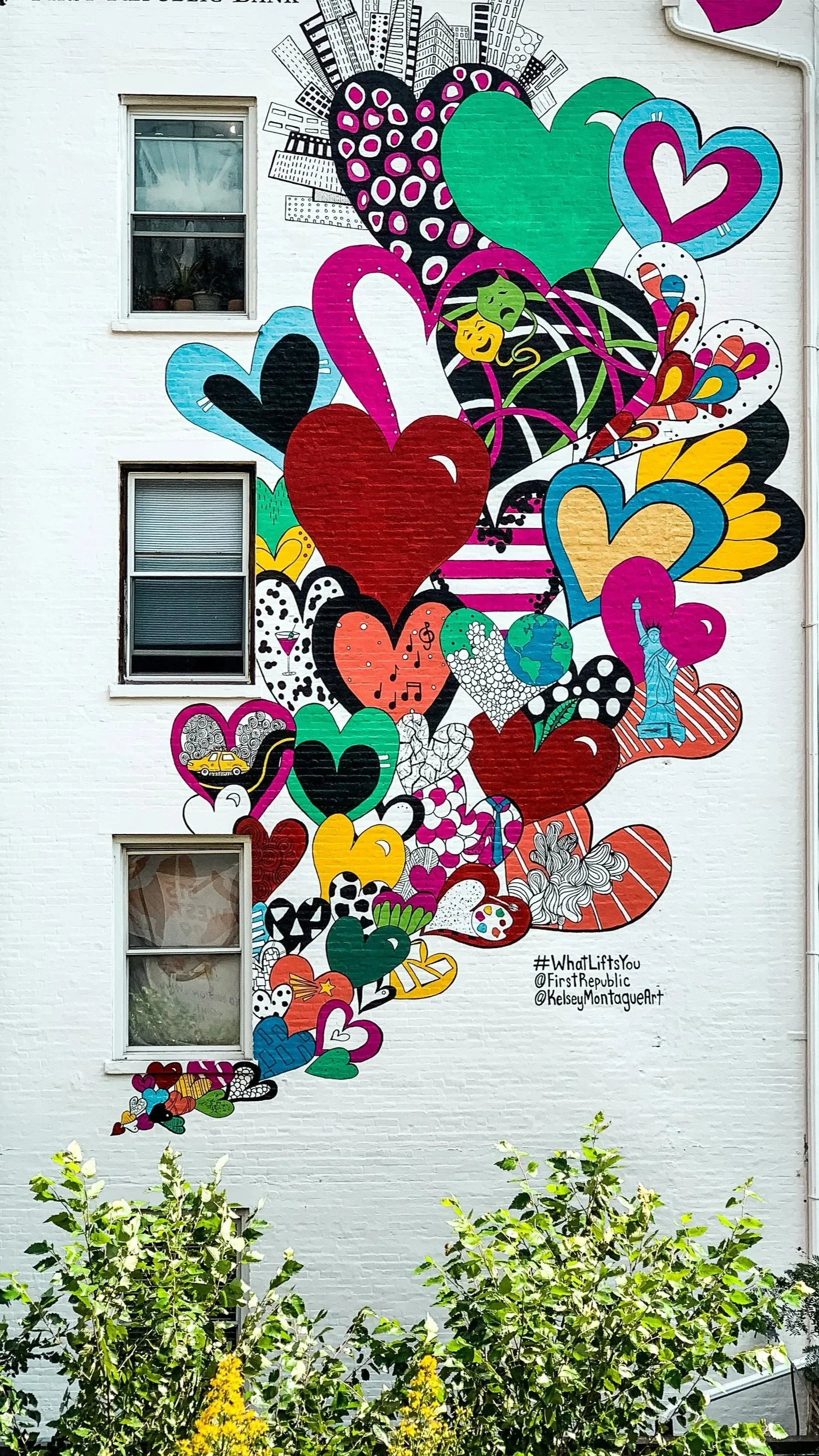 Colorful mural of various hearts, symbols, and images on a white brick building, including the Statue of Liberty, Earth, music notes, and cityscape, with greenery at the bottom.
