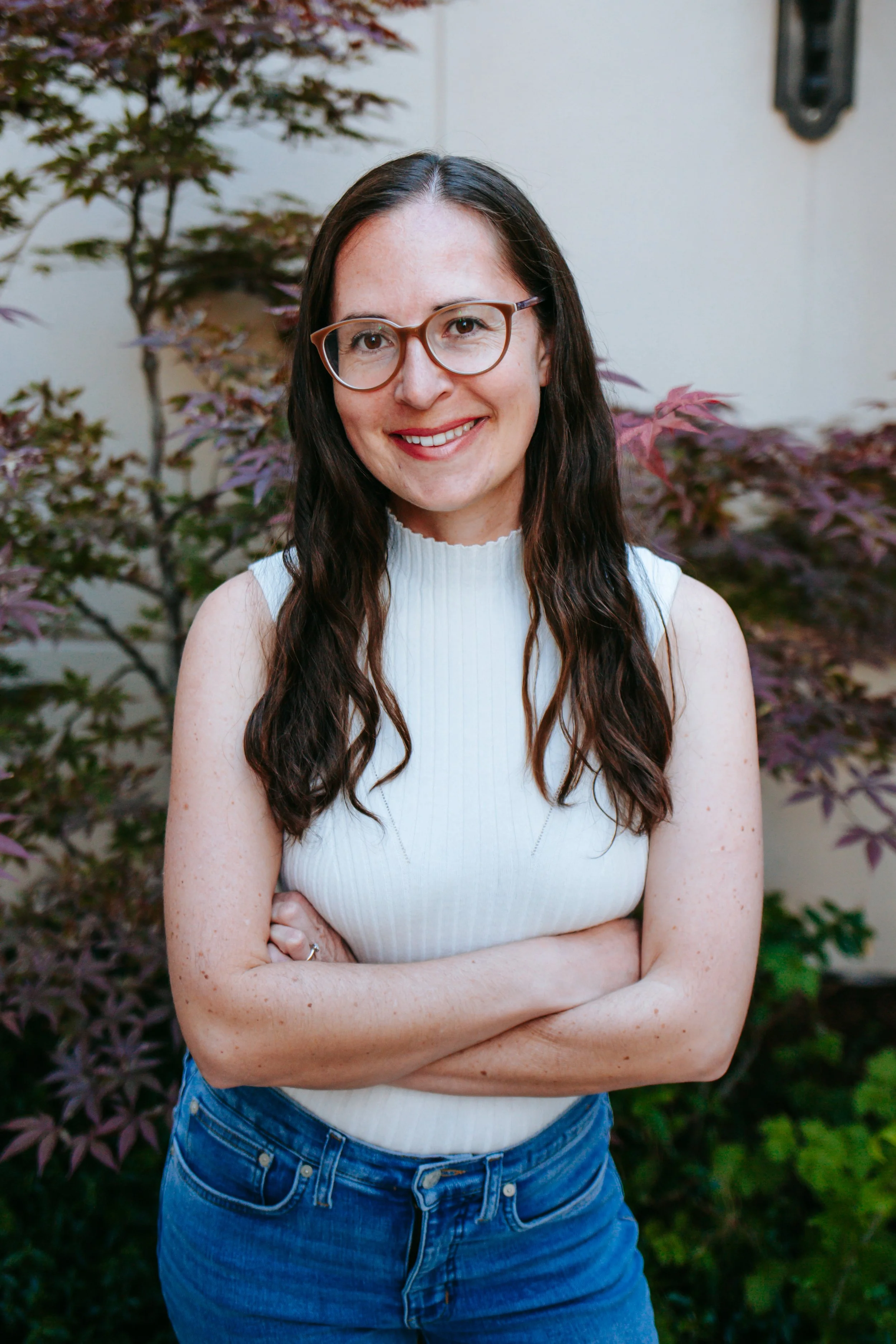 A woman with long dark hair, glasses, and a striped shirt smiling outdoors, with green foliage in the background.