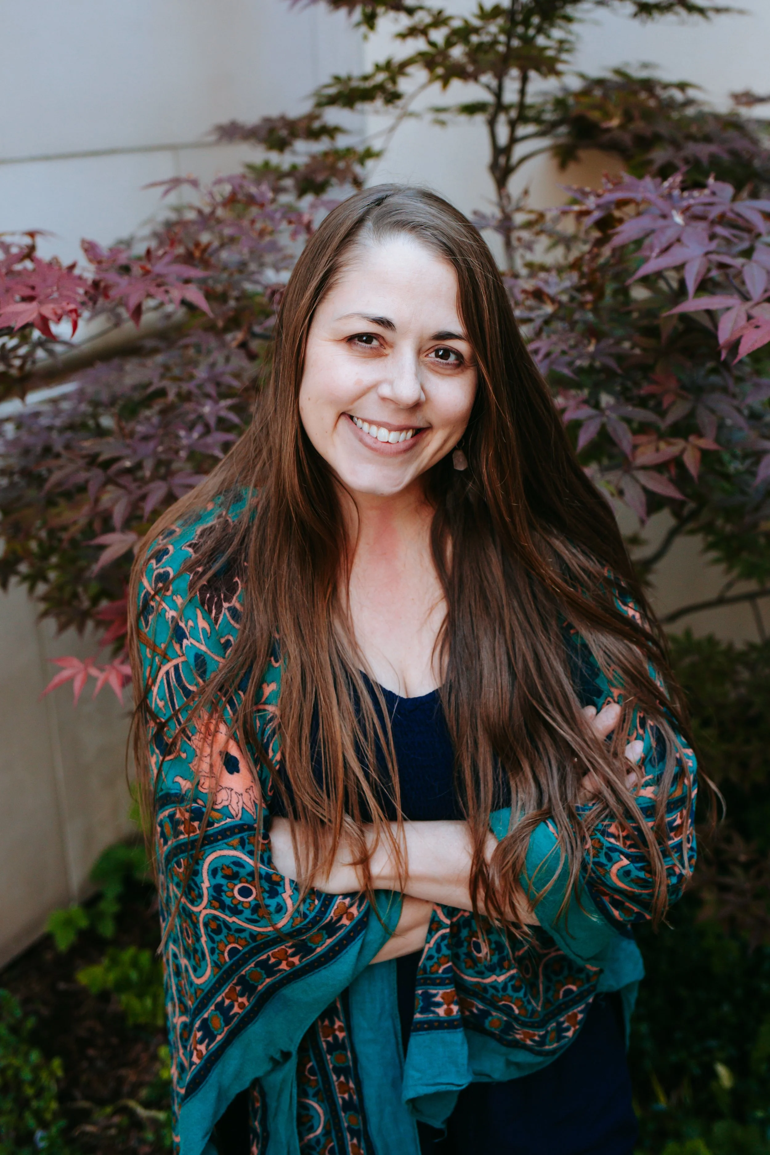 A woman with long brown hair smiling outdoors, wearing a red dress and a floral shawl, standing next to a wooden railing with lush green trees in the background.