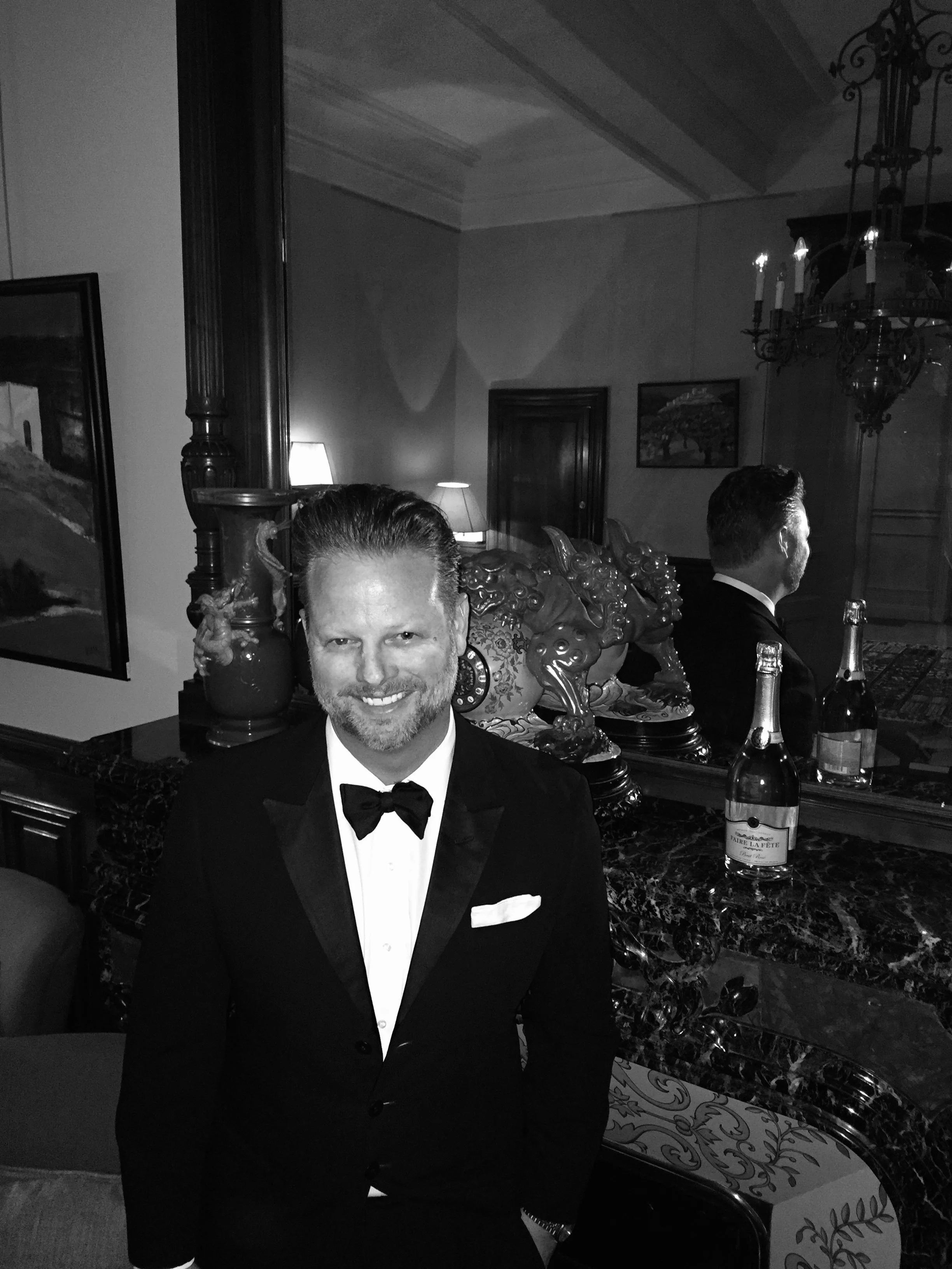 Peter Baedeker in a tuxedo in front of a fireplace with a bottle of Champagne on the mantle. 
