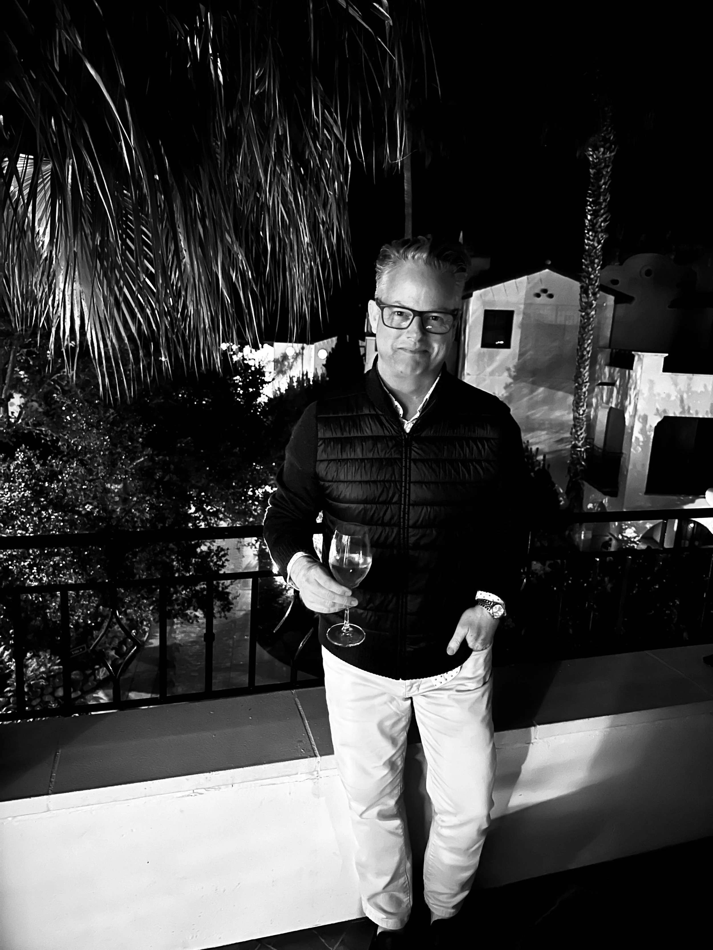 Black and white photo of Peter Baedeker standing with a black sweater and holding a wine glass, palm tree to his right and Spanish style building behind him