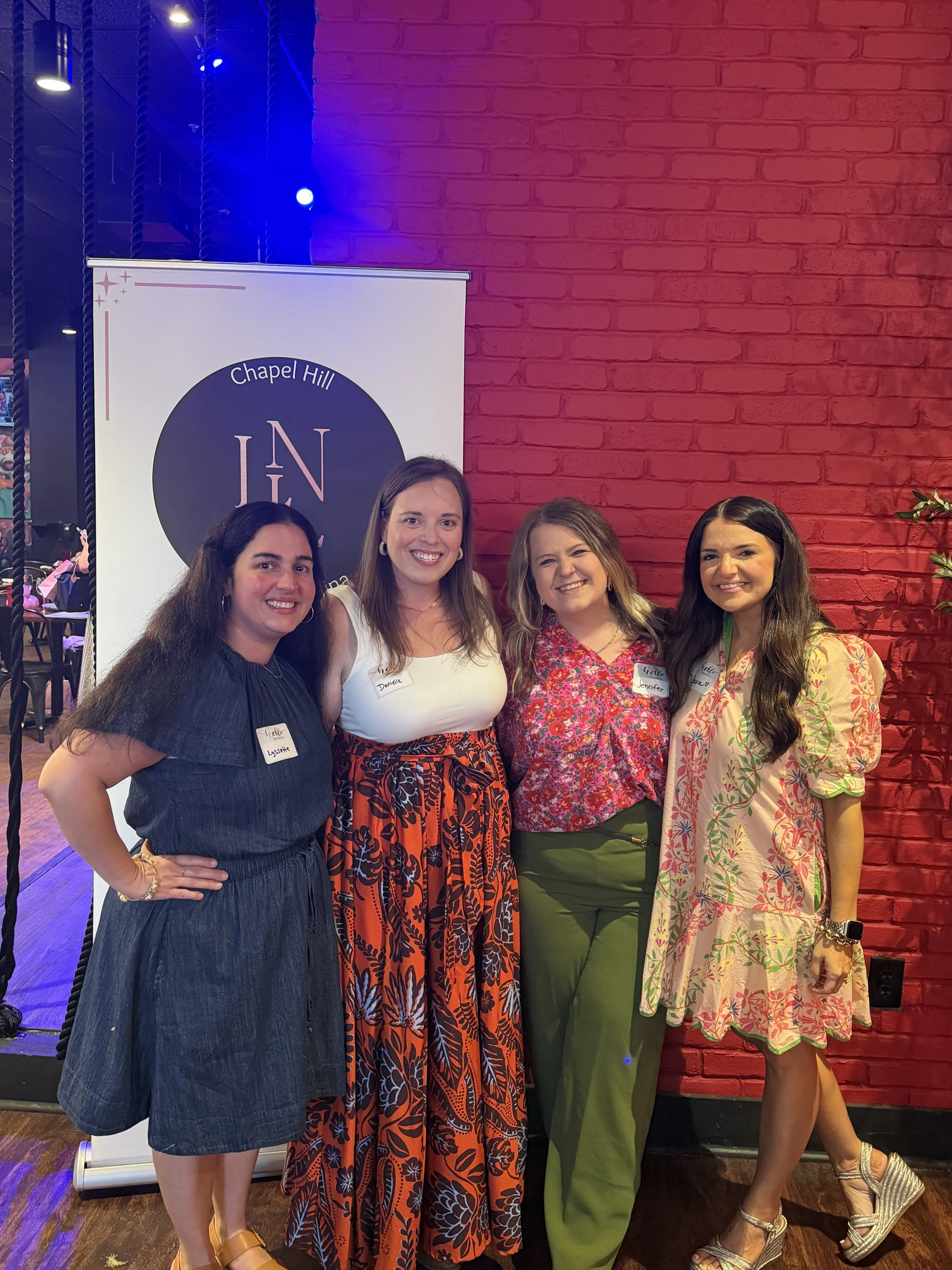 Group of four women standing in front of a red brick wall at an indoor event, smiling for a photo. The woman on the far left wears a dark blue dress with a name tag that says 'Lesette'. The second woman from the left has long brown hair, wearing a white top and a colorful long skirt. The third woman has wavy hair, wearing a pink floral blouse and green pants. The woman on the far right has long dark hair, wearing a light-colored dress with a floral pattern and high heels. Behind them is a sign that says 'Chapel Hill' and 'LN'.