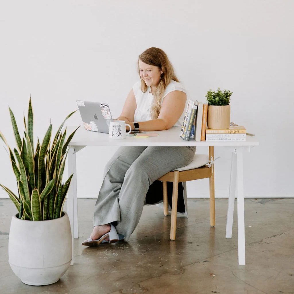 A woman sitting at a white desk working on a laptop, with a potted plant, books, and a coffee mug in a bright, minimalistic office space.