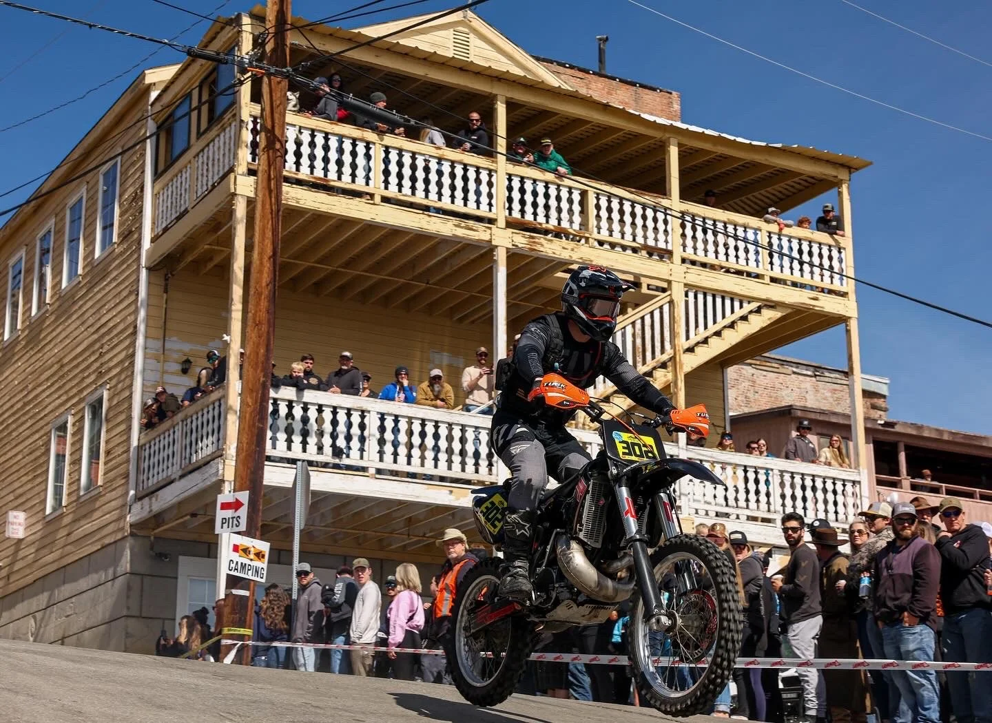 A motocross rider wearing black riding gear and helmet, on a black motorcycle with orange accents, racing past a crowd of spectators gathered on the street and watching from balconies of a wood building during a daytime event.